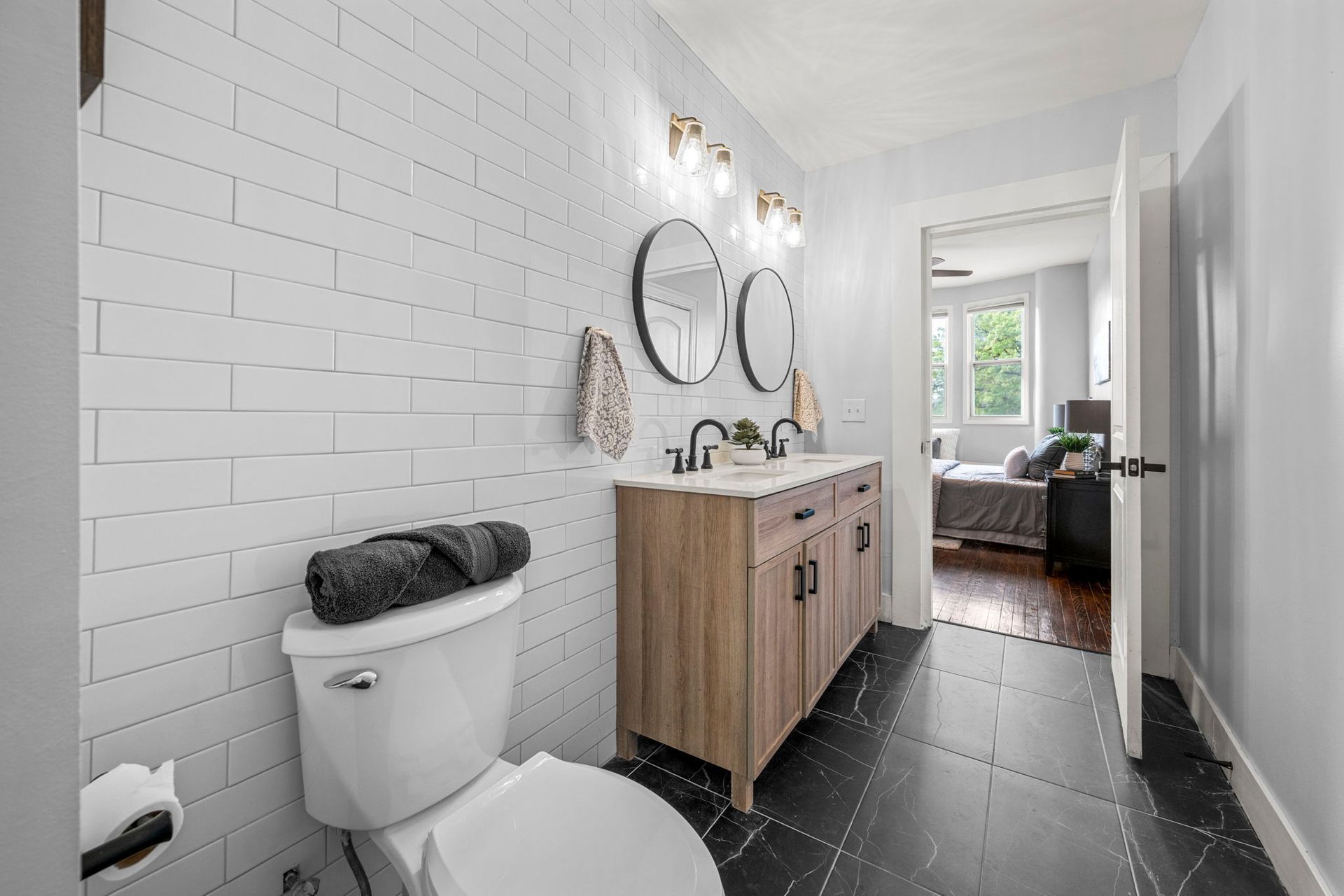 Bathroom with white brick-like tile, wood vanity with two sinks, black floor tile, and open doorway to bedroom.