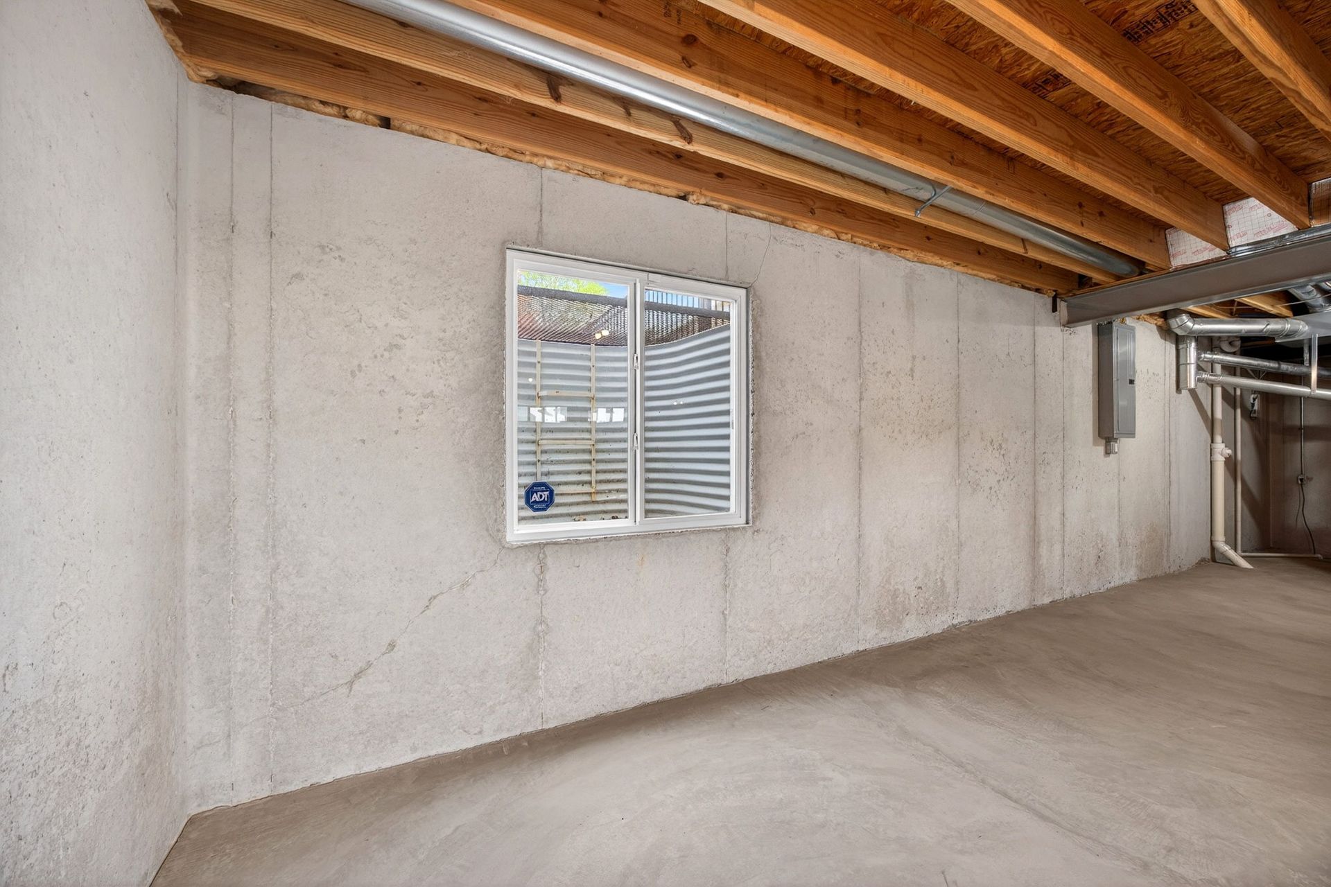 Unfinished basement with concrete walls, a small rectangular window, and exposed wooden ceiling joists.