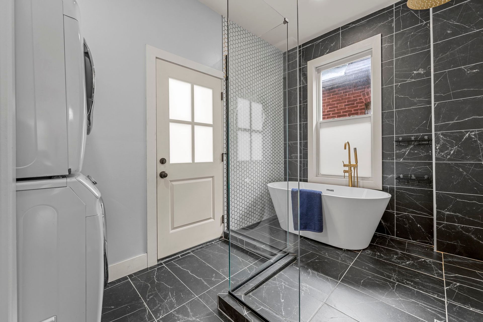 Modern bathroom with stacked washer/dryer, black tiled shower, freestanding tub, and exterior door.
