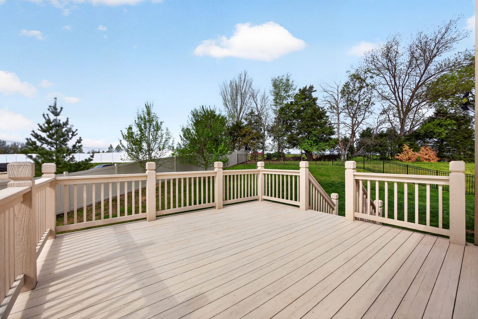 A light-colored wooden deck with matching railings overlooks a grassy backyard with trees under a sunny blue sky.