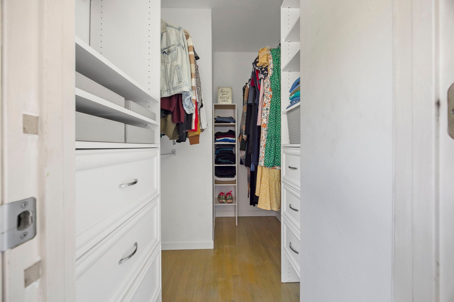 Walk-in closet with white shelves, drawers, and clothes hanging. Wooden floor.