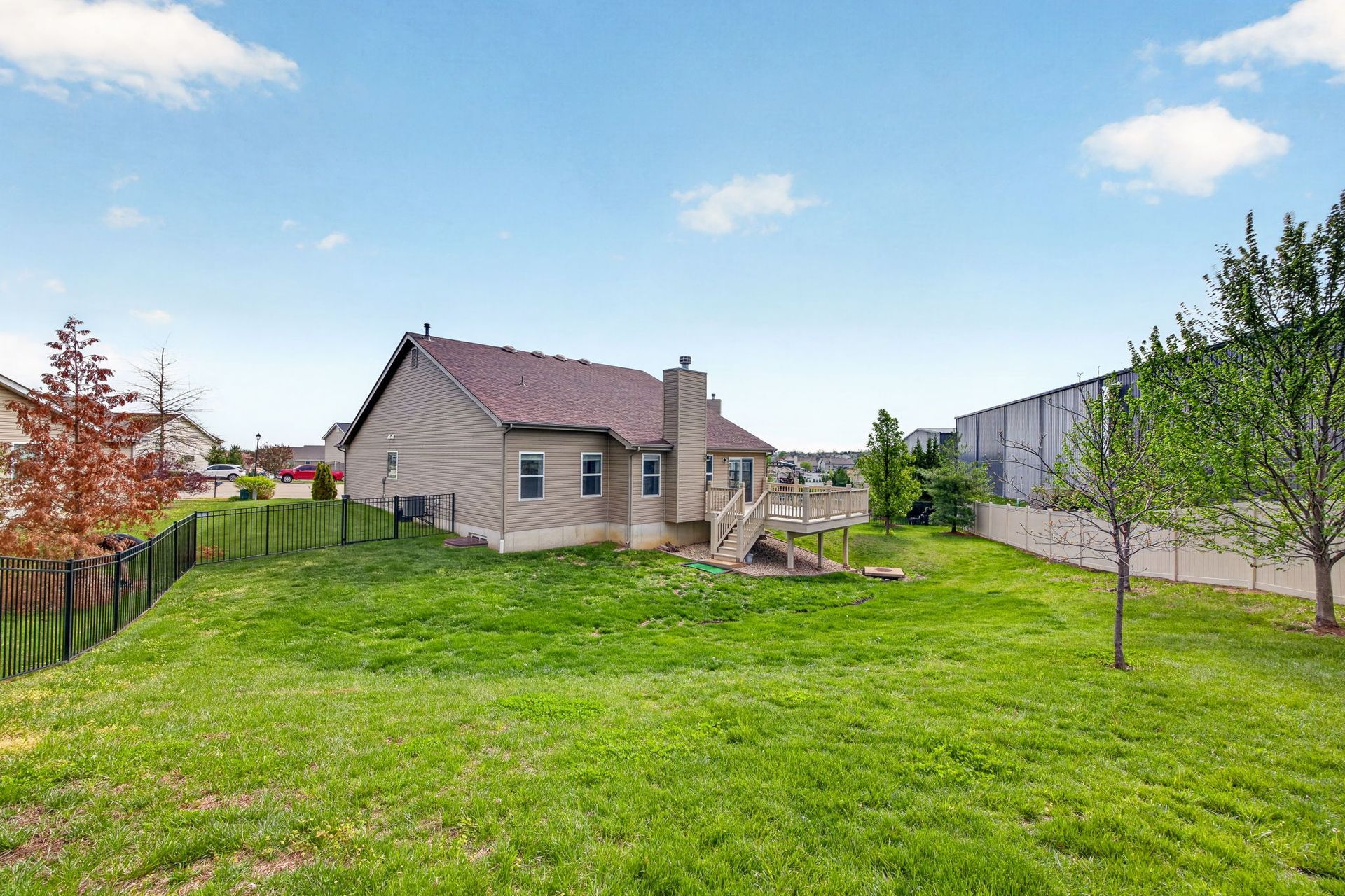 Backyard view of a tan, one-story house with a chimney and small wooden deck, set against a green lawn and cloudy blue sky.