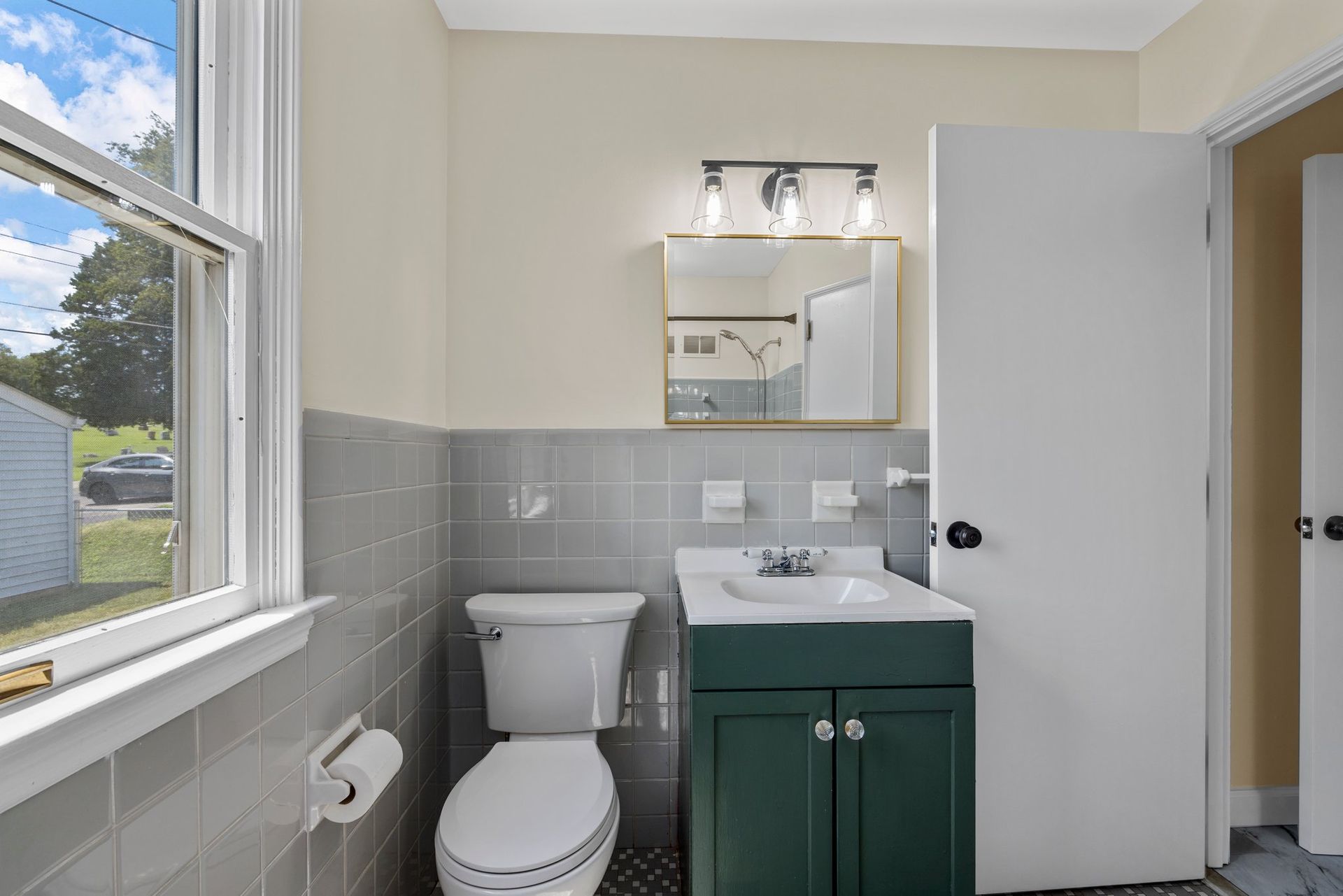A bathroom with a toilet , sink , mirror and window.