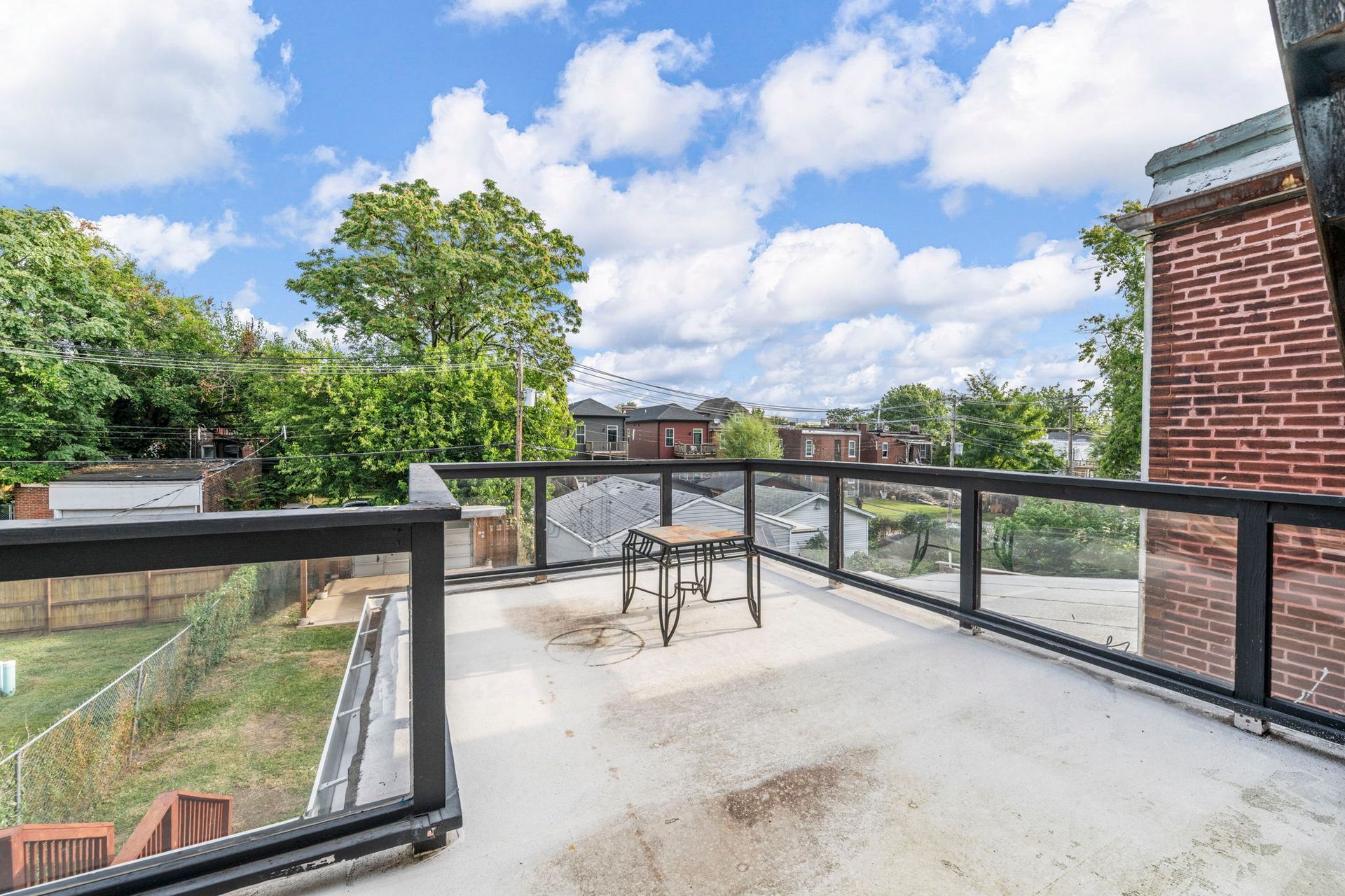Rooftop patio with glass railing overlooking backyard, trees, and other buildings, under a partly cloudy sky.