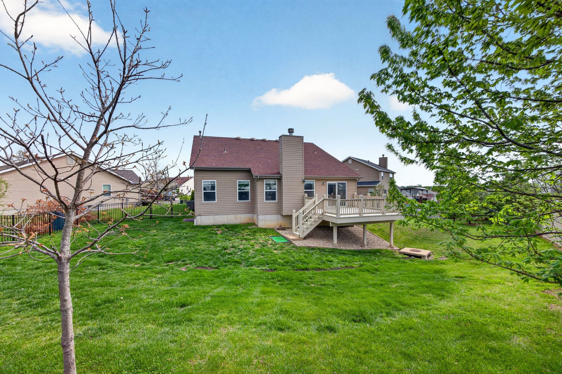 A rear view of a two-story beige house with a wooden deck, a chimney, and a grassy backyard under a partly cloudy sky.