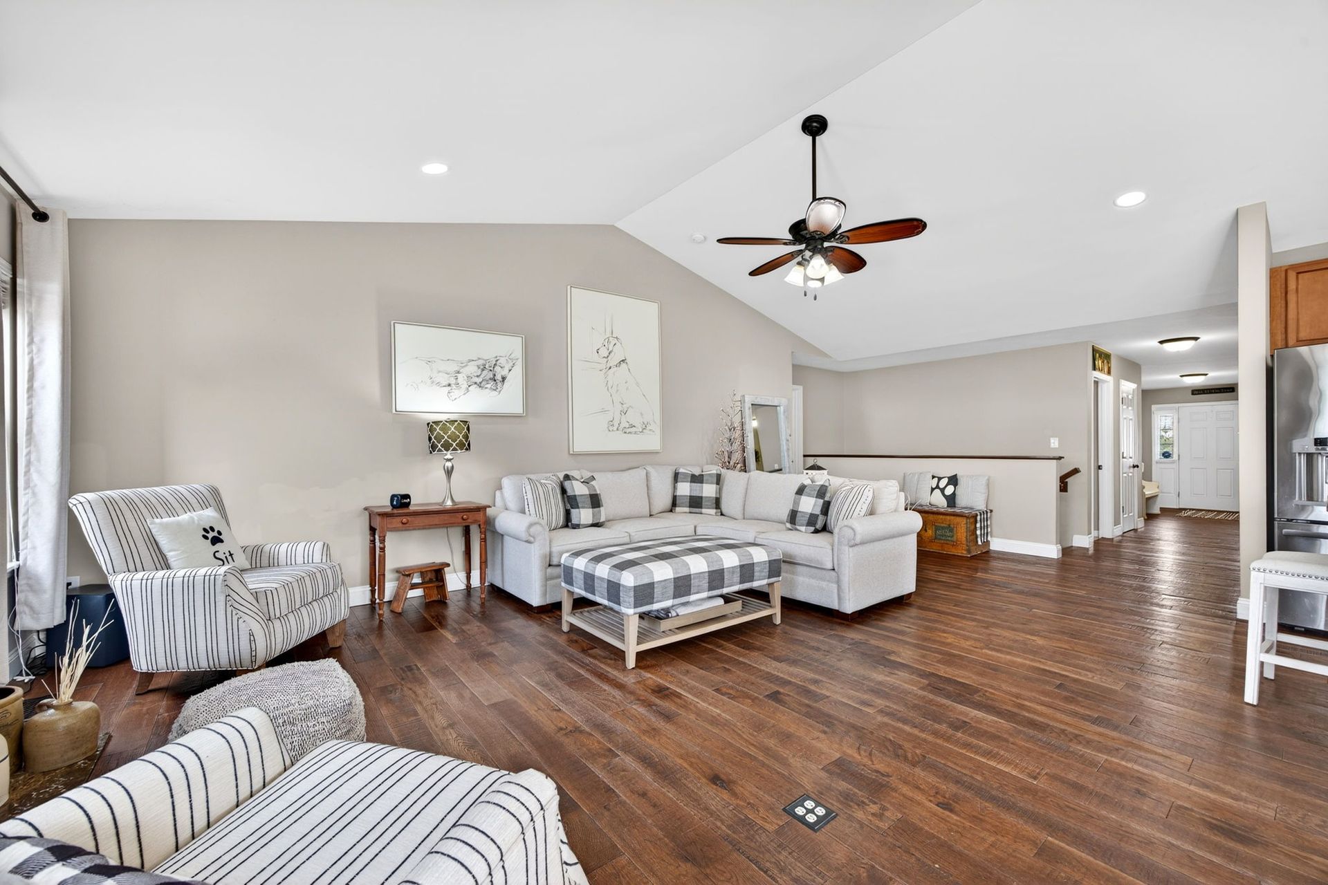 A spacious living room with hardwood floors, a white sectional sofa, a plaid ottoman, and an armchair under a ceiling fan.