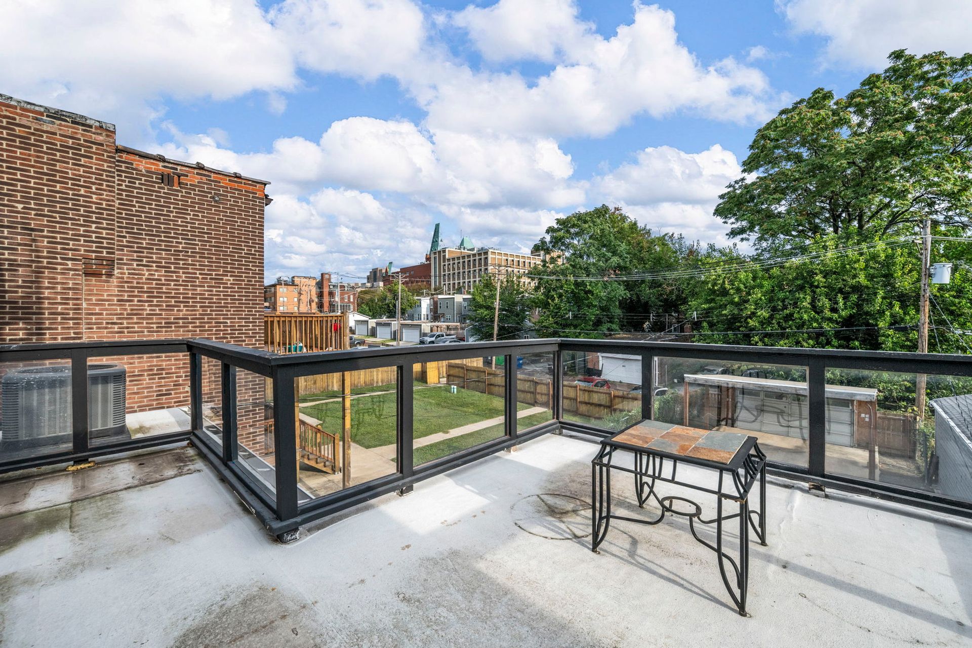 Rooftop patio with glass railing, small table, overlooking backyard, buildings, and trees. Blue sky with clouds.