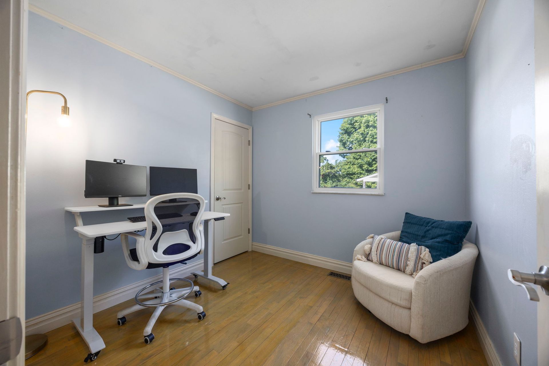A light blue office with a desk, chair, monitor, armchair, and window; hardwood floor.
