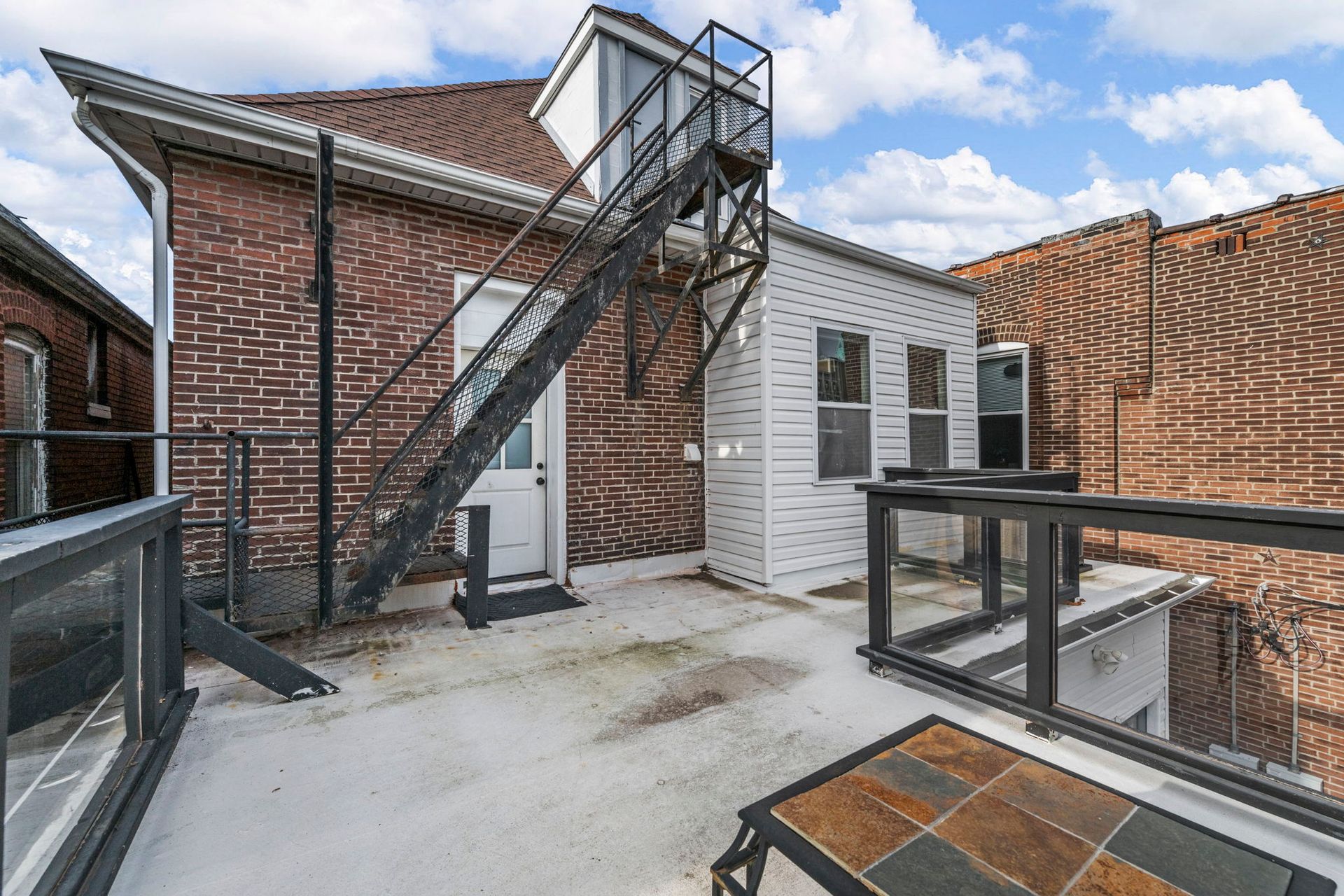 Rooftop patio with brick building, metal fire escape, white walls, and glass railings.