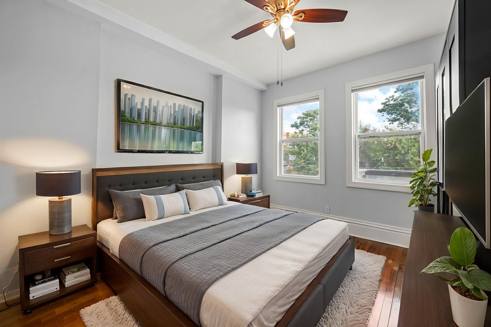 Bedroom with king bed, two windows, TV, and artwork. Grey walls, wooden floors, and a ceiling fan.