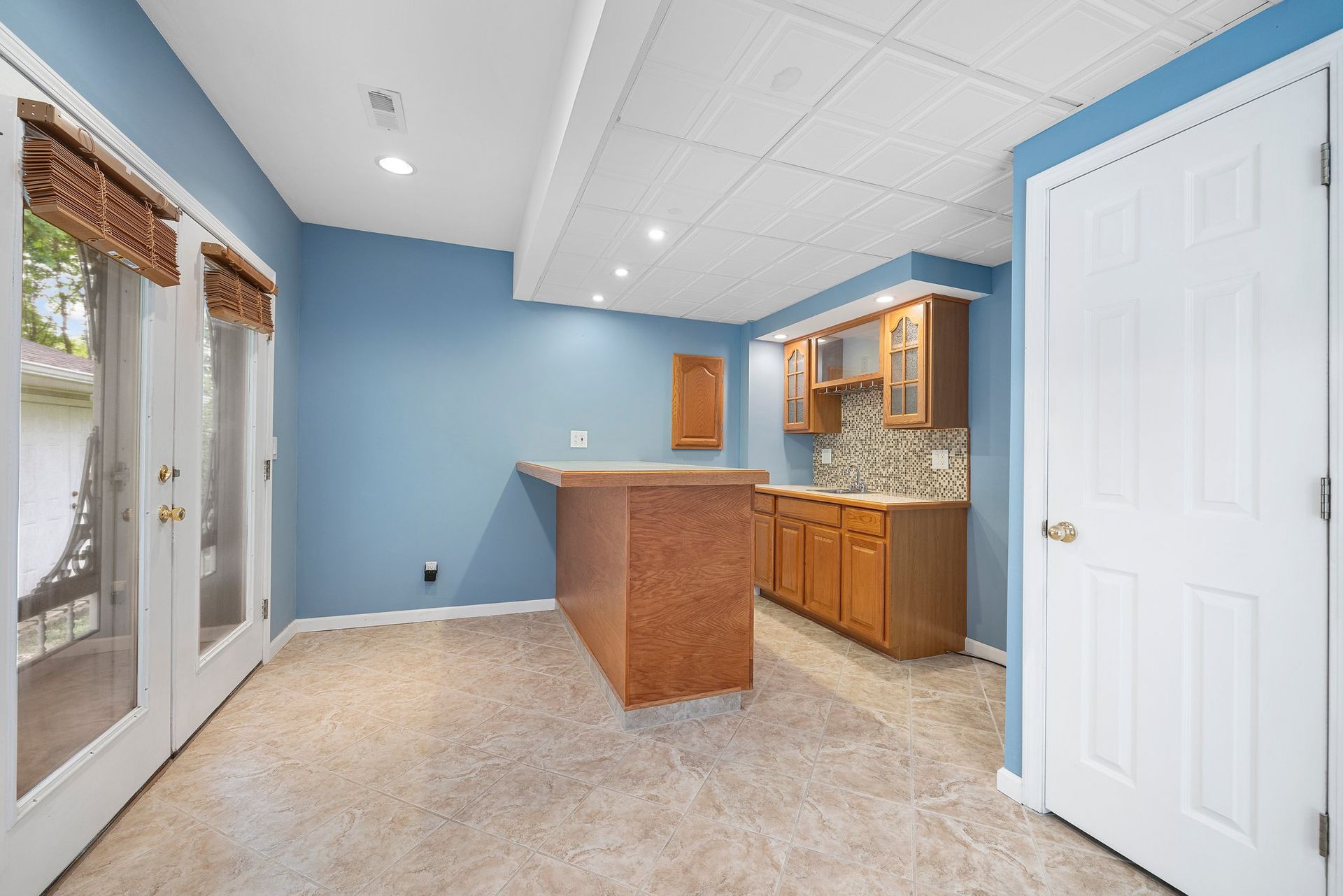 A basement with blue walls , wooden cabinets , a bar and sliding glass doors.