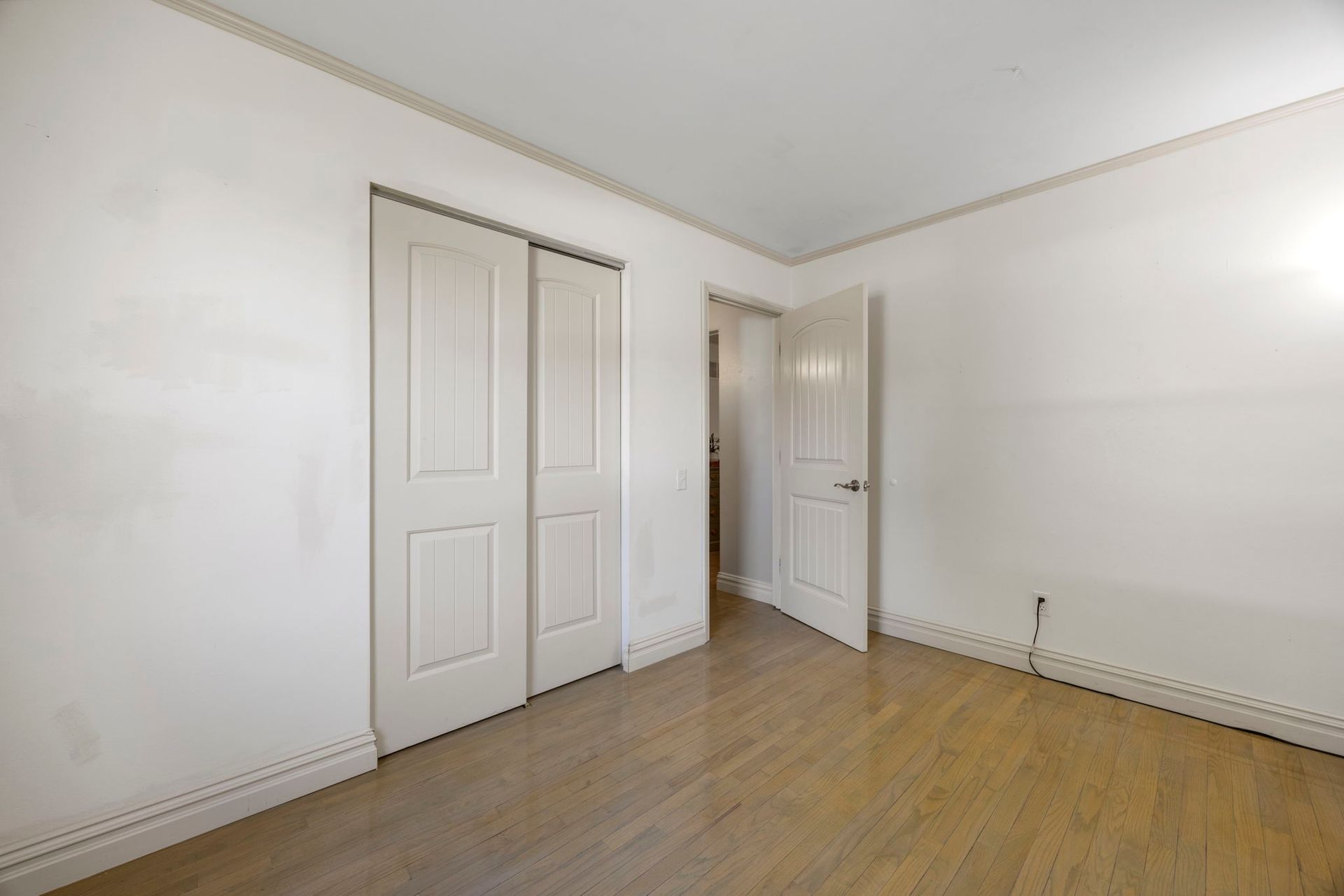 Empty bedroom with hardwood floors, white walls, closet, and open doorway.