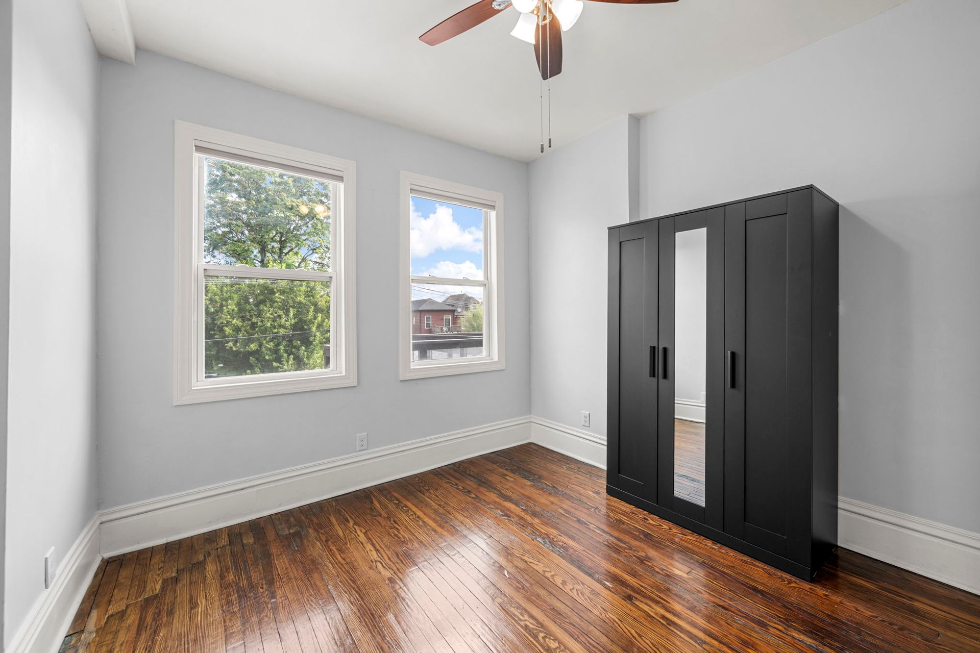 Empty bedroom with hardwood floors, two windows, black wardrobe, and a ceiling fan.