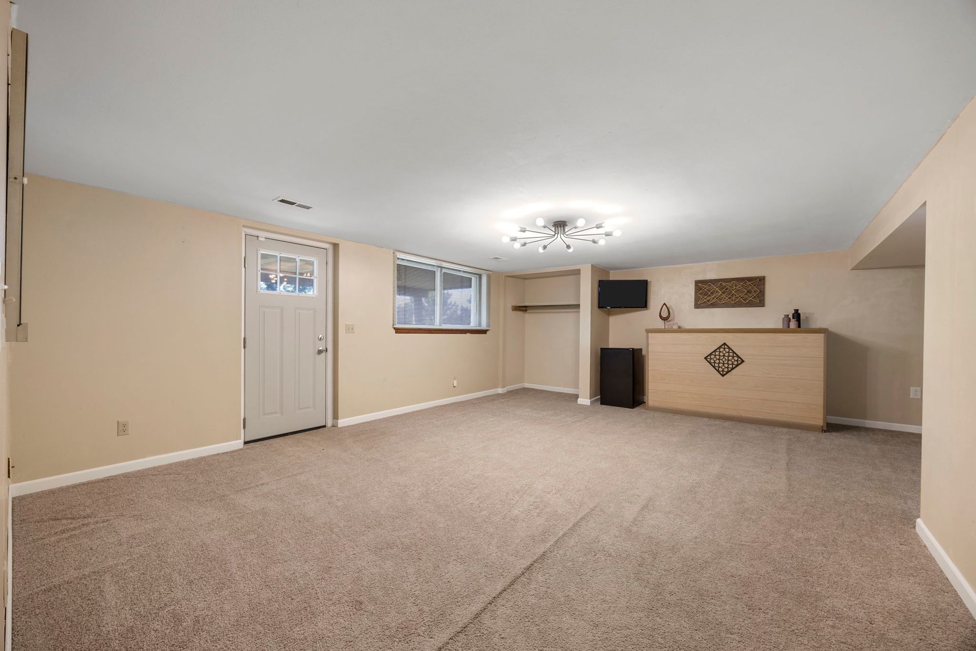 A basement room with carpet, a bar, a window, and a door. Beige walls and light fixture.