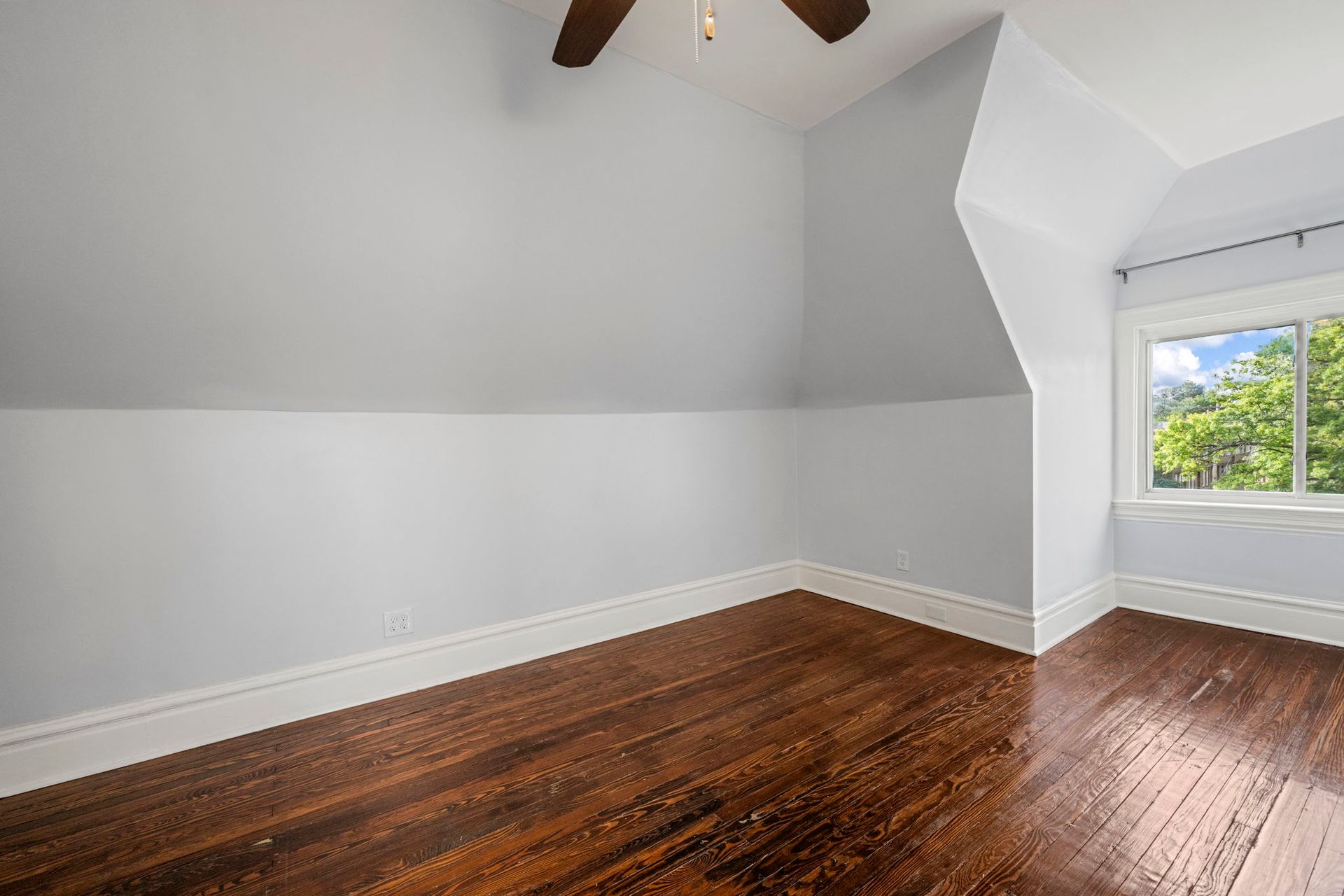 Empty room with light blue walls, wood floor, and a window with a view of trees.