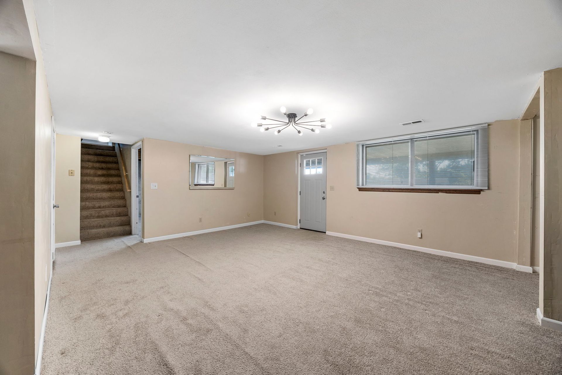 Empty beige basement room with carpet, door, window, and staircase.