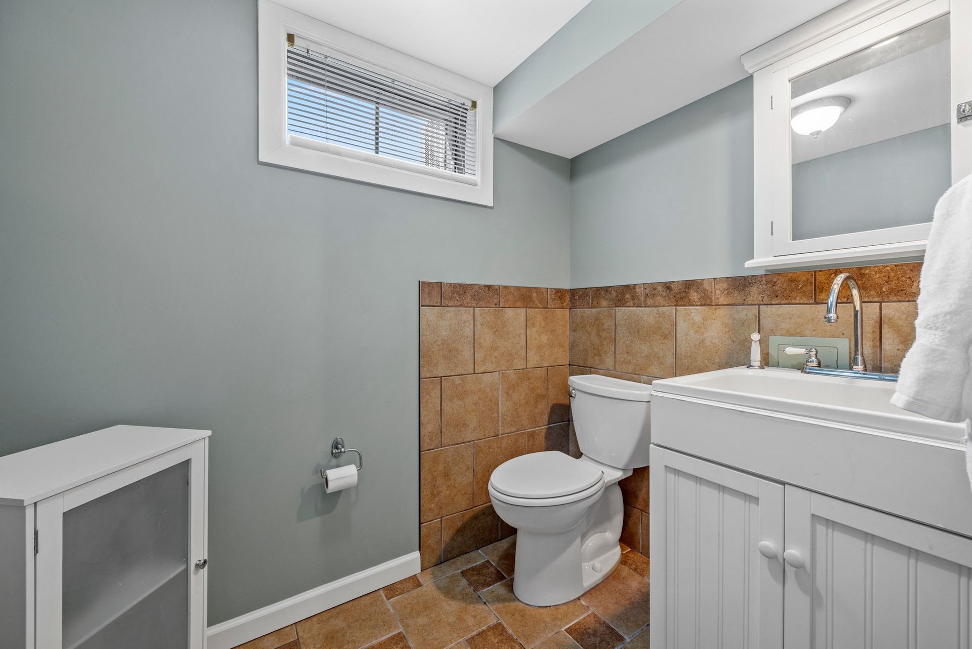 A bathroom with a toilet , sink , mirror and window.