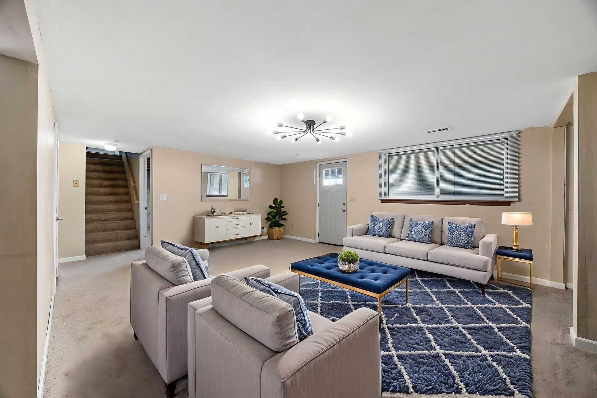Living room: beige walls, carpet, blue rug, white couch with blue pillows, armchairs, coffee table, and a staircase.