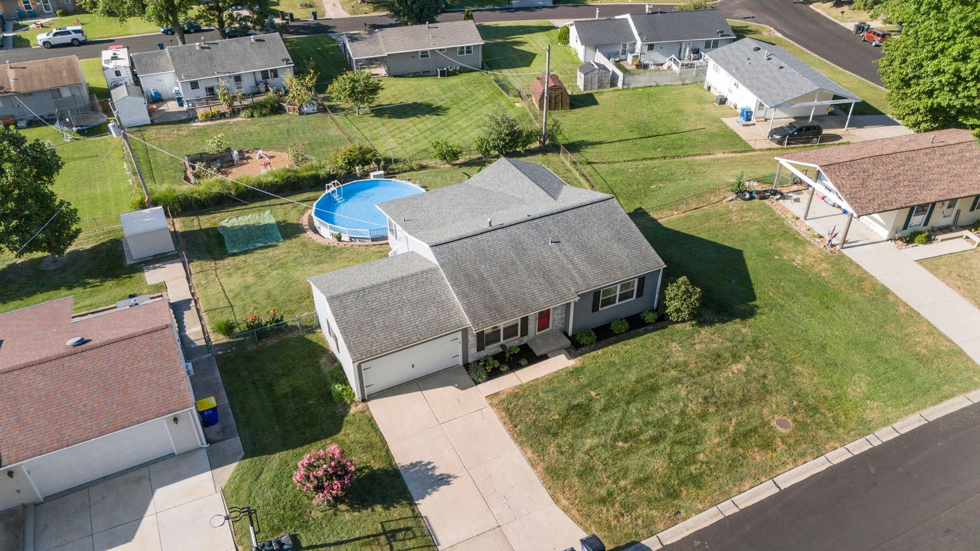 Aerial view of a single-family house with gray siding, a pool, and a driveway in a suburban neighborhood.
