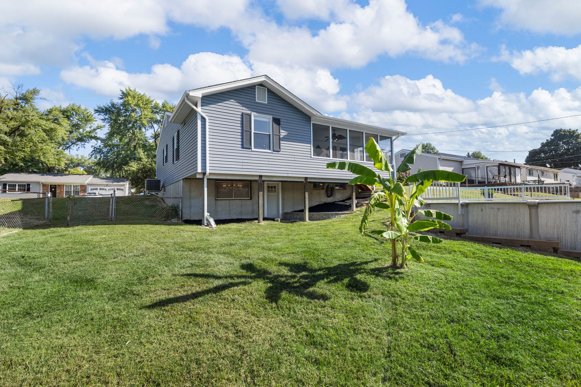 Backyard view of a light-blue house with a screened porch, banana tree, and grassy lawn under a blue sky.
