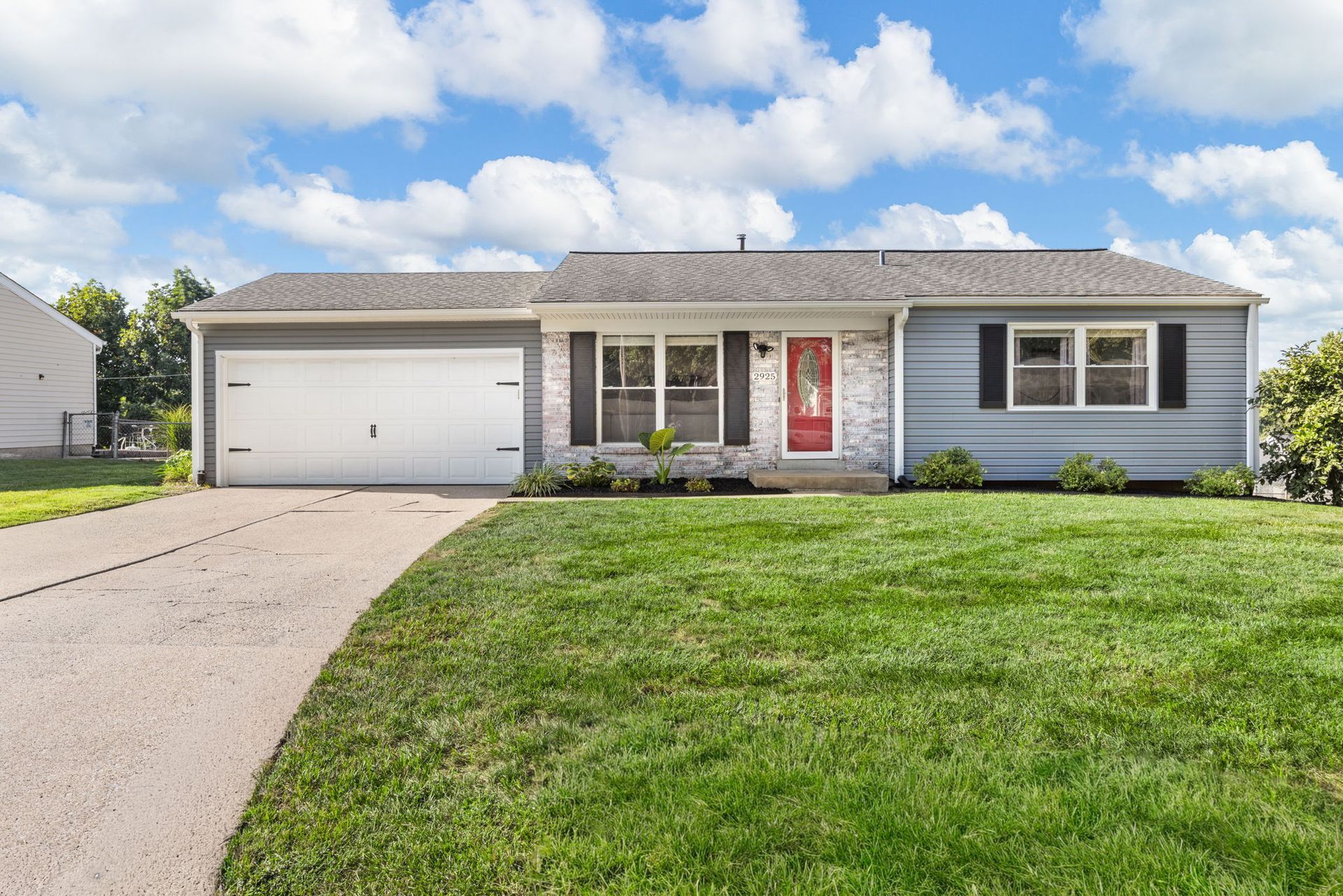 Ranch-style home with gray siding, white garage door, brick facade, red door, and green lawn.