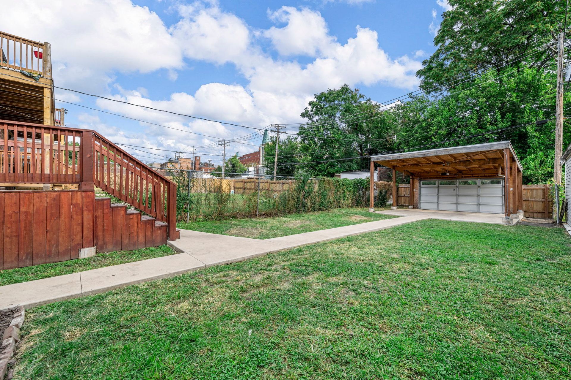 Backyard with wooden deck, grassy lawn, concrete path, and a carport on a sunny day.