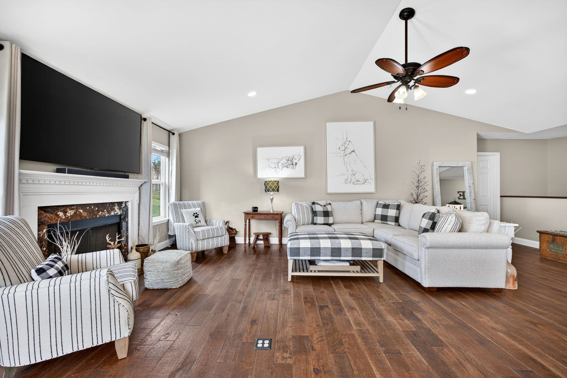 A bright living room with dark wood floors, a fireplace, a large TV, patterned armchairs, and a beige L-shaped sofa.