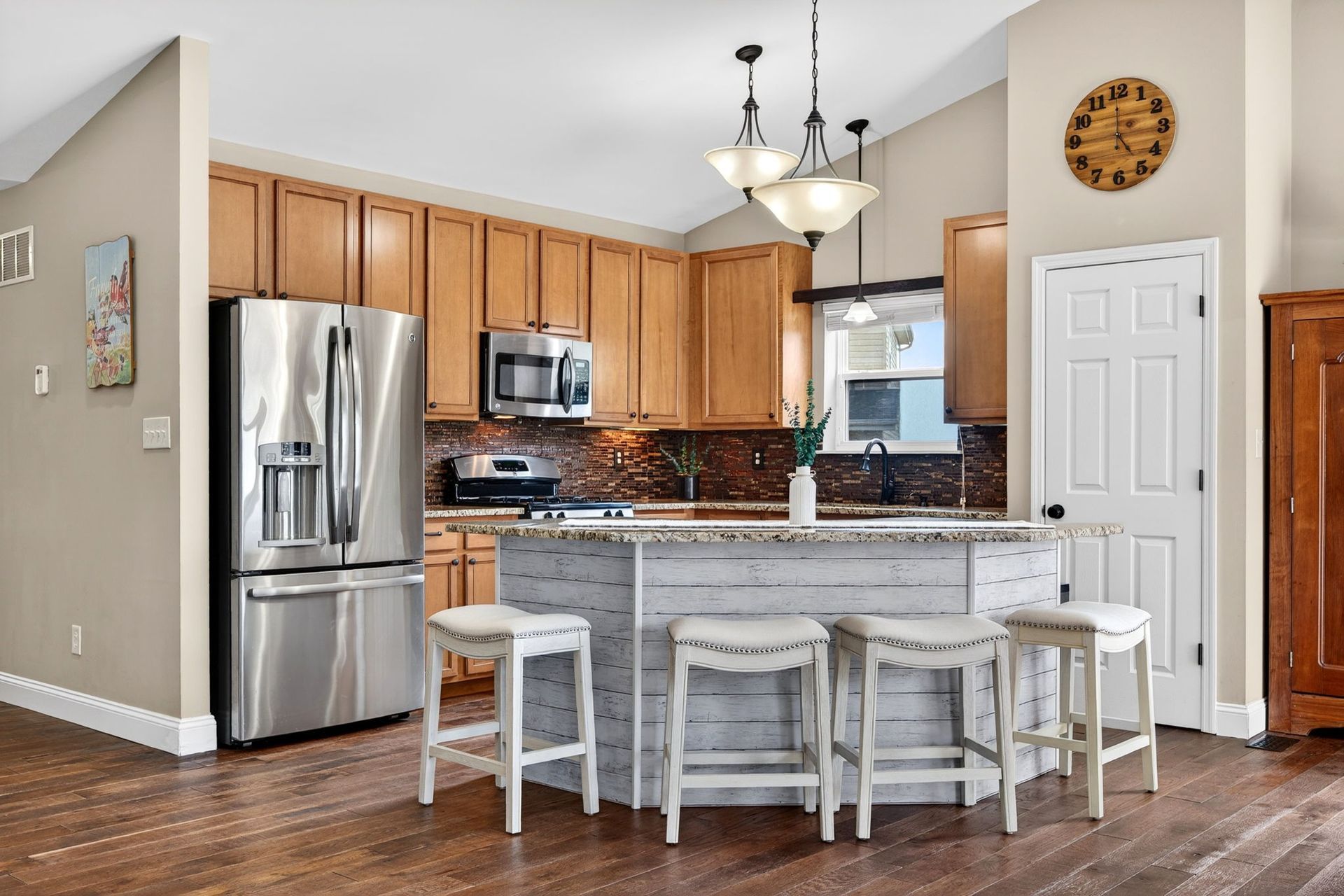 A modern kitchen with wooden cabinets, stainless steel appliances, a white island with four stools, and a round wall clock.