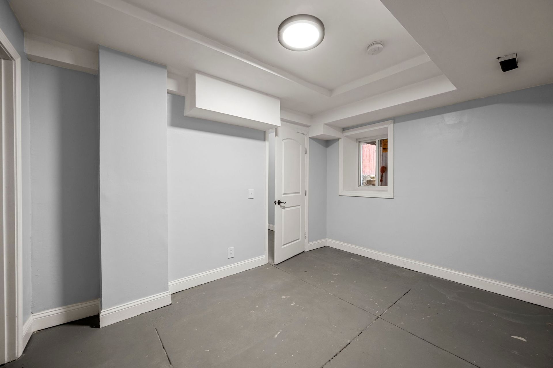 Empty room with light blue walls, white trim, and gray floor mats; a closed door, a window, and a ceiling light are visible.