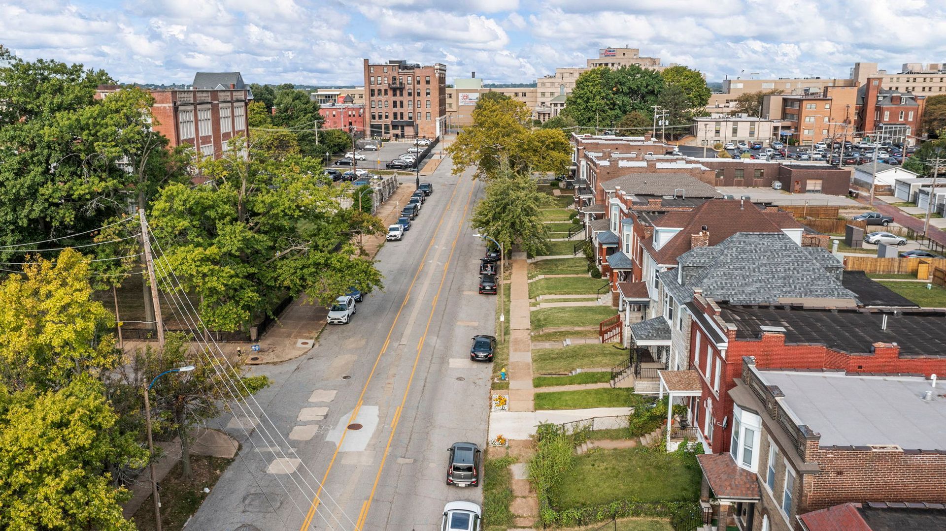 Aerial view of a residential street lined with trees, parked cars, and brick buildings under a cloudy sky.