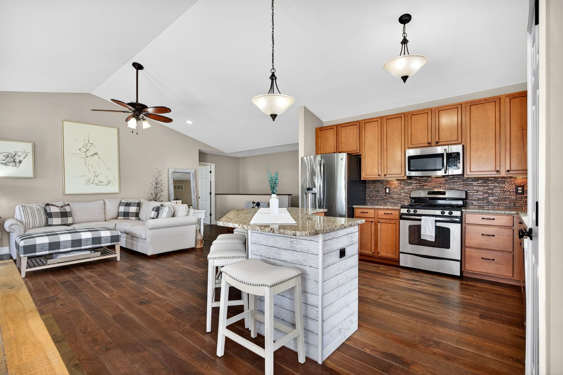 A modern open-concept kitchen with wood cabinets and an island, adjacent to a living area with a sofa and vaulted ceiling.