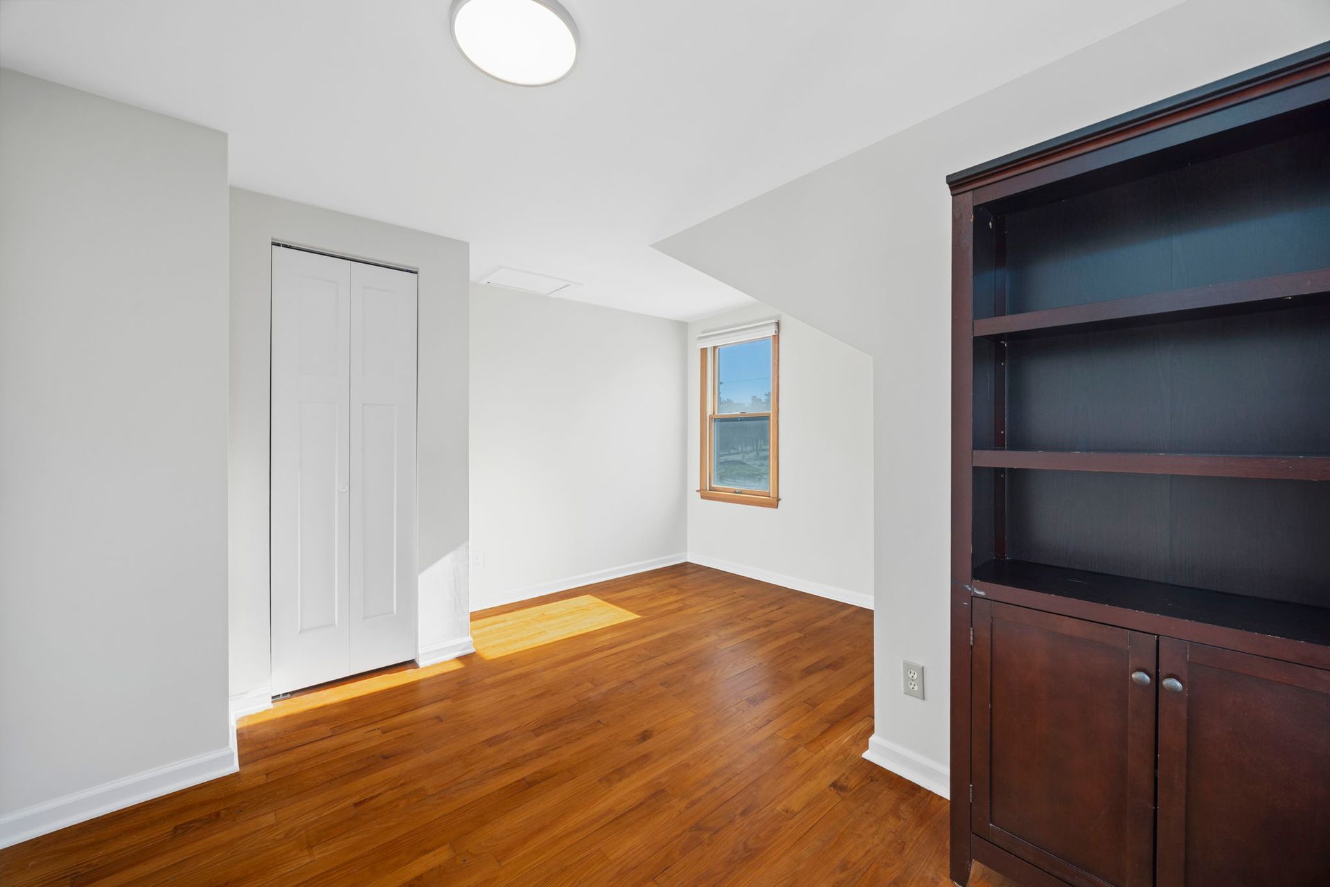 An empty room with hardwood floors , a bookshelf and a window.