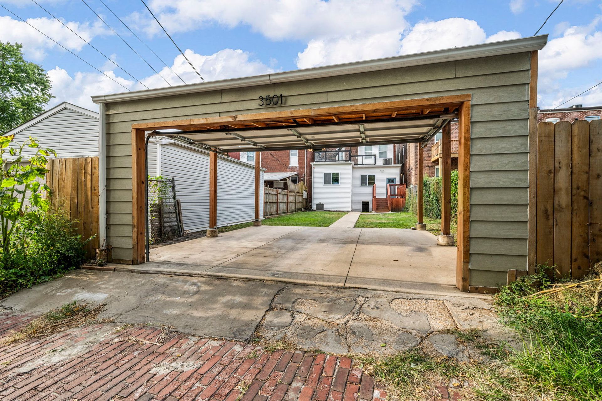 Open-air carport with concrete driveway, leading to a backyard with houses and green space.