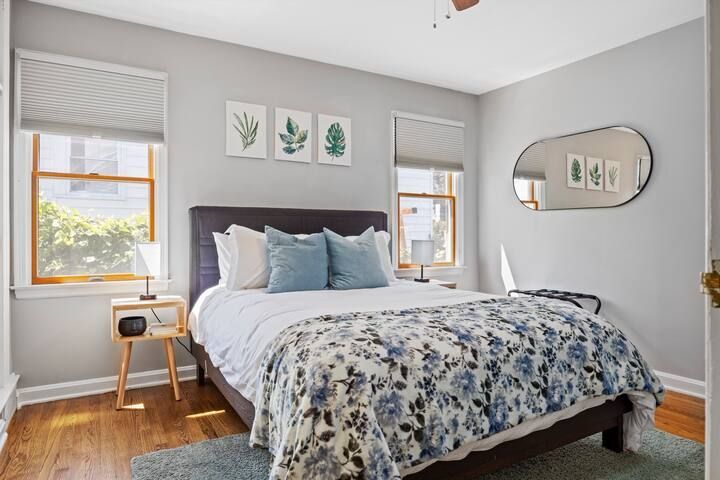 A bedroom with a bed , nightstand , mirror and two windows.