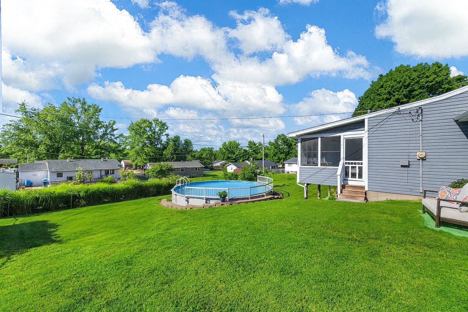 Green backyard with an above-ground pool. Blue sky and white clouds, a gray house with a screened porch.