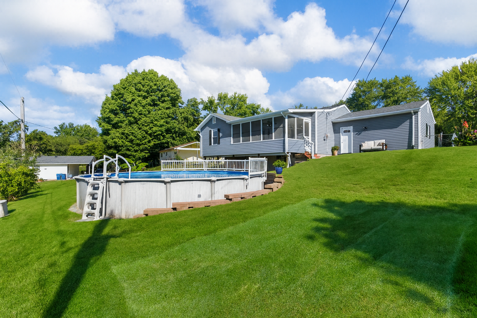 Backyard with above-ground pool, sloped green lawn, and a blue house on a sunny day.