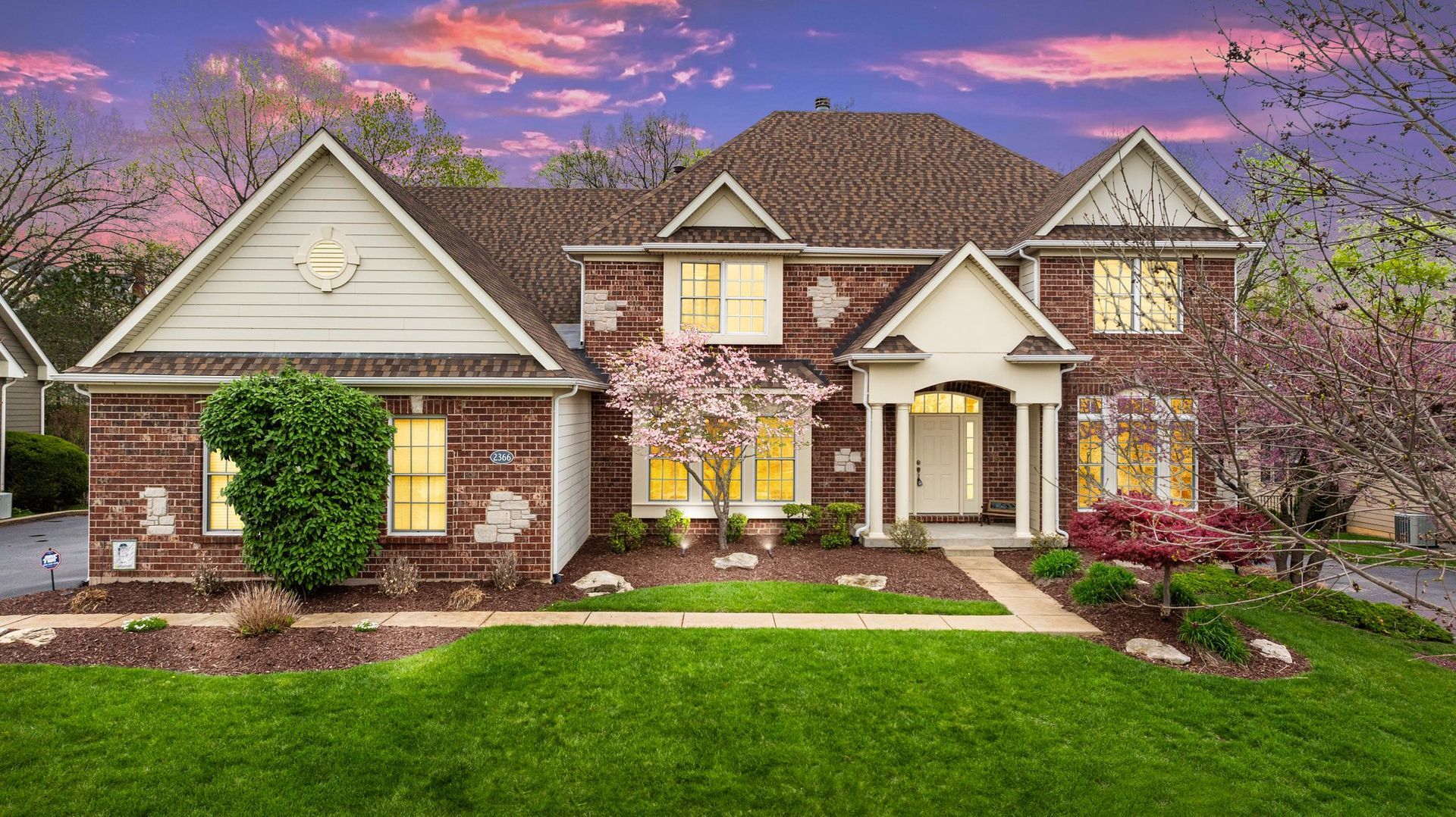 A large brick house with a lush green lawn and a sunset in the background.