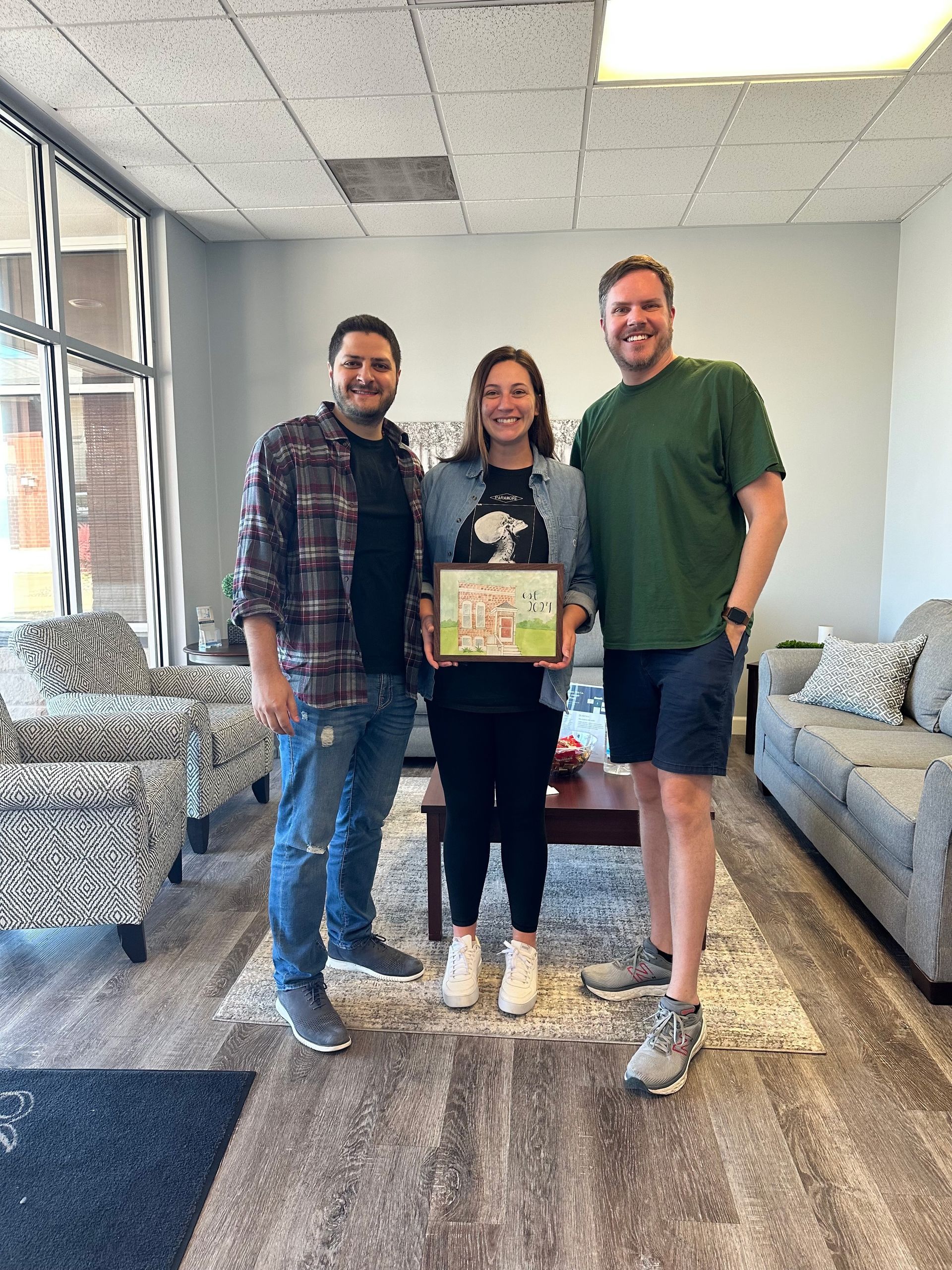 Three people are standing next to each other in a living room holding a picture.