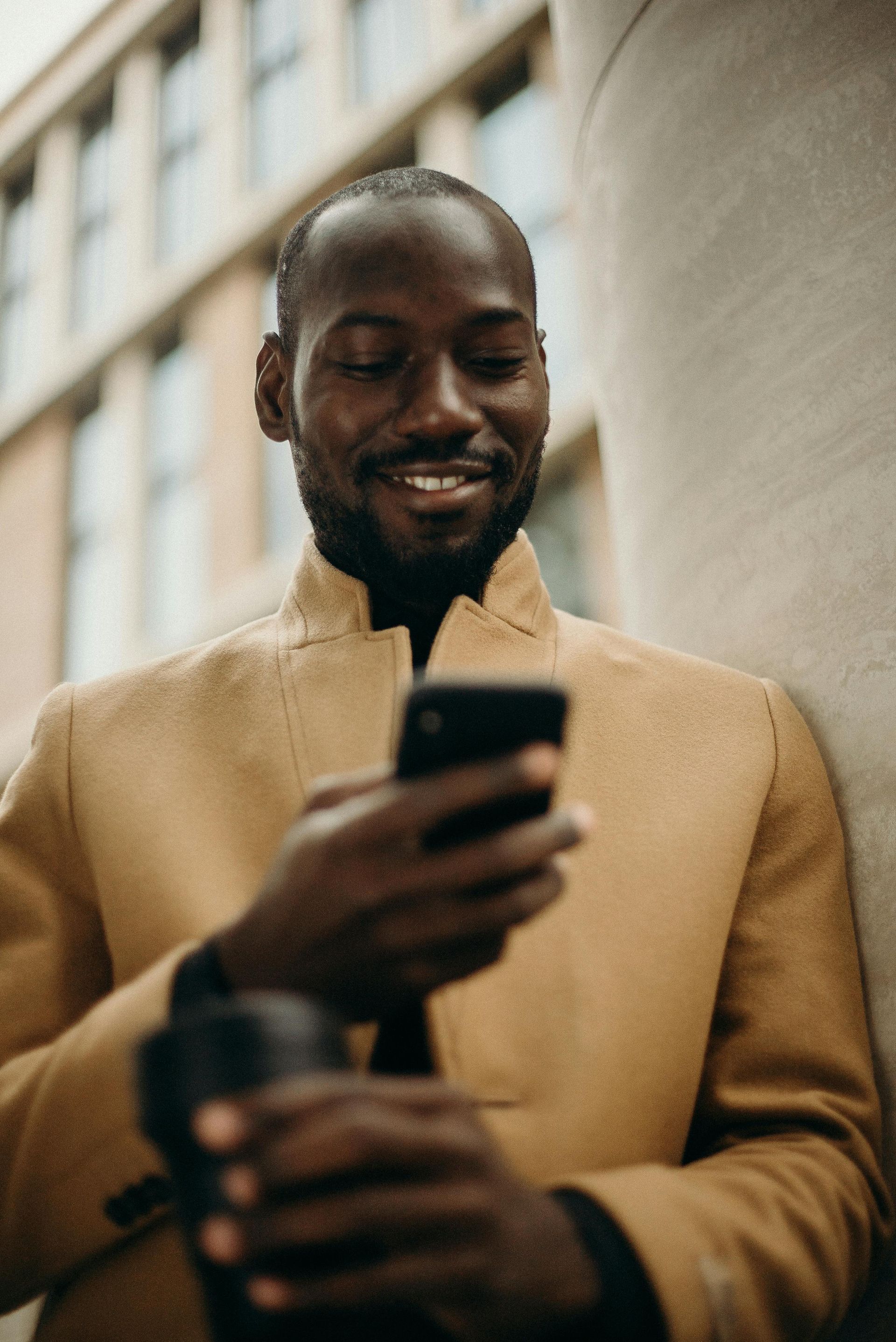 A man is leaning against a wall while looking at his cell phone.