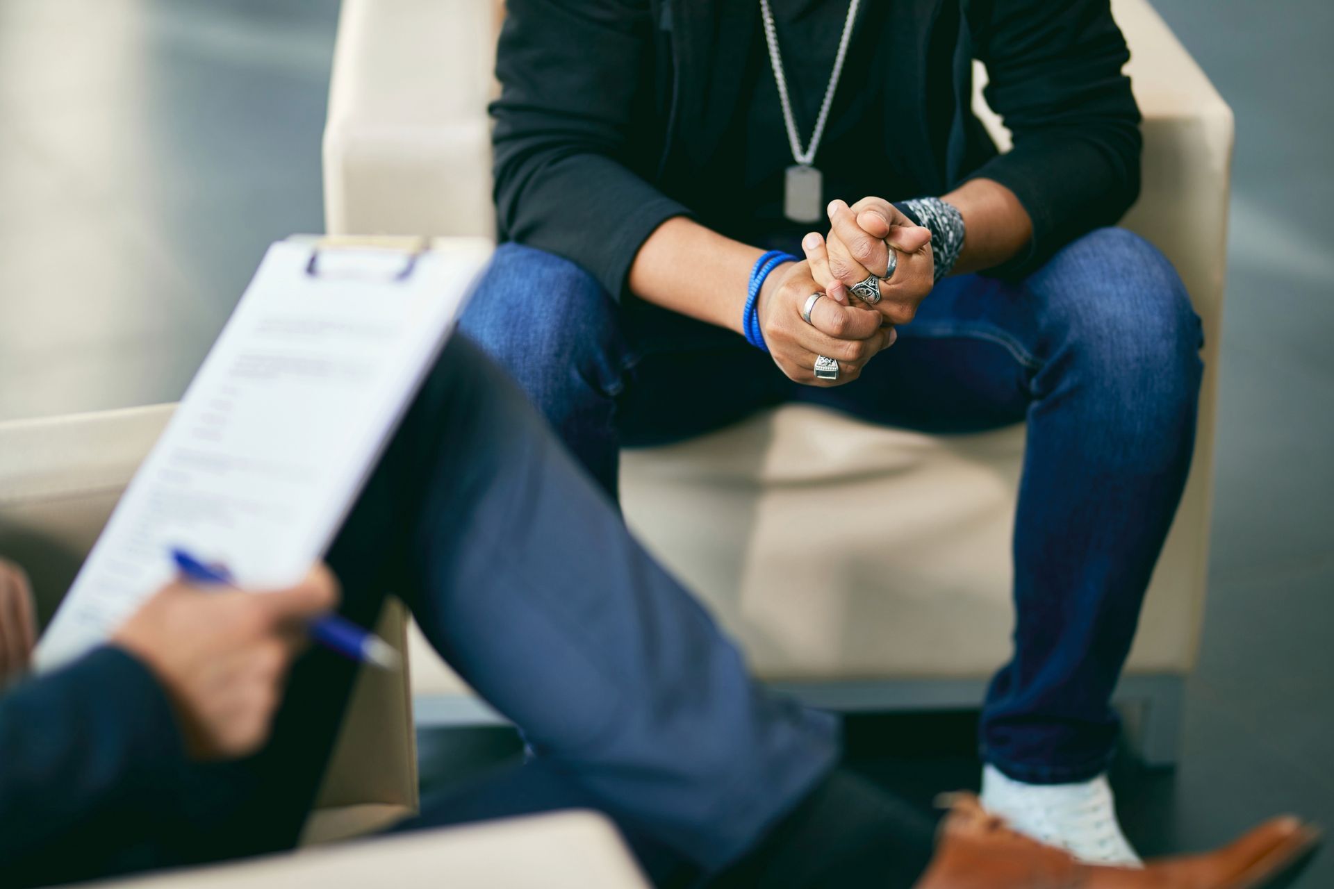 Person in jeans sits in an armchair, hands clasped. Someone else holds a clipboard and pen.