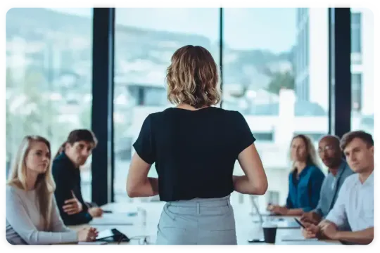 A woman is giving a presentation to a group of people sitting around a table.