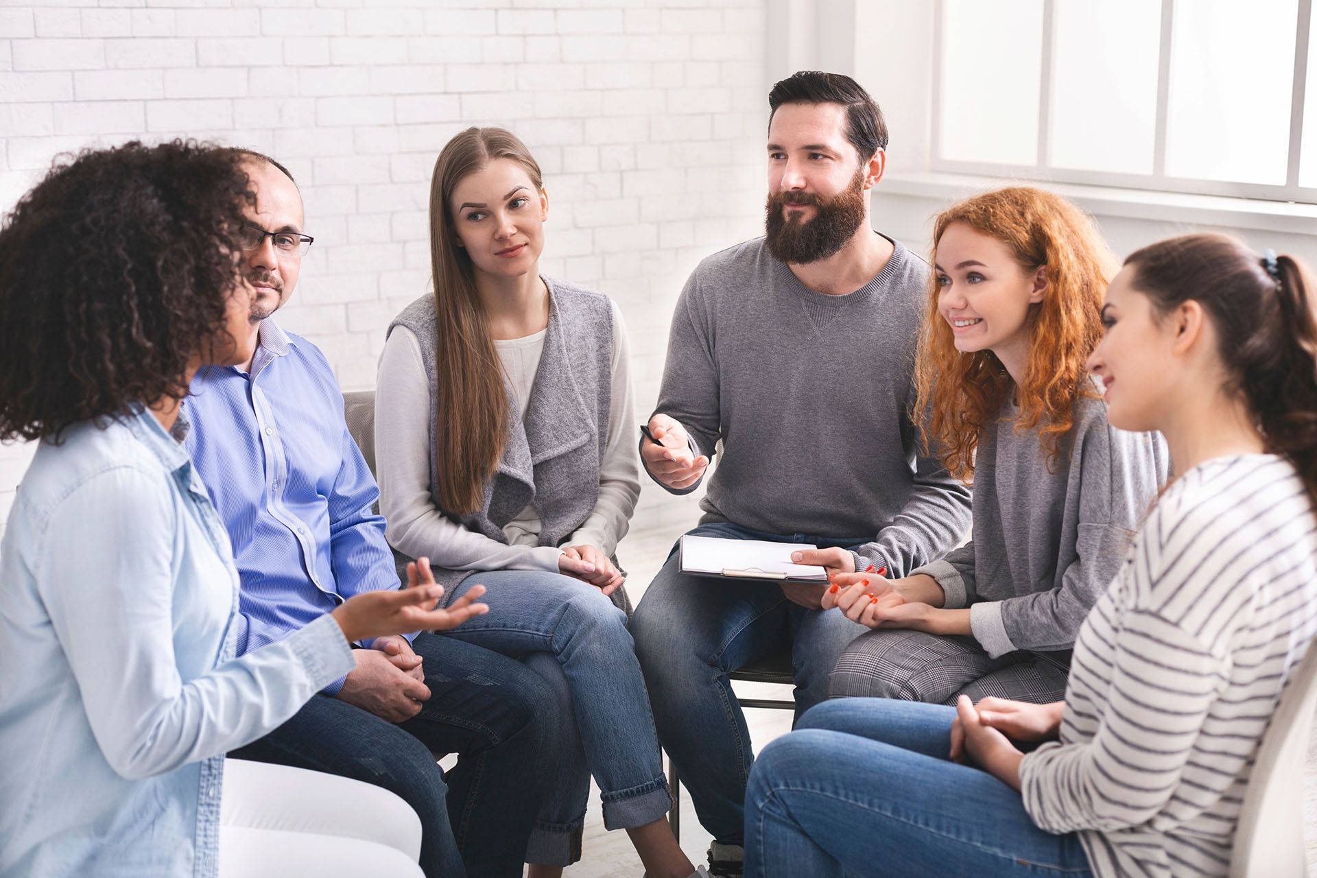 Group of people in a circle, conversing in a light-filled room. They are diverse in age and race.