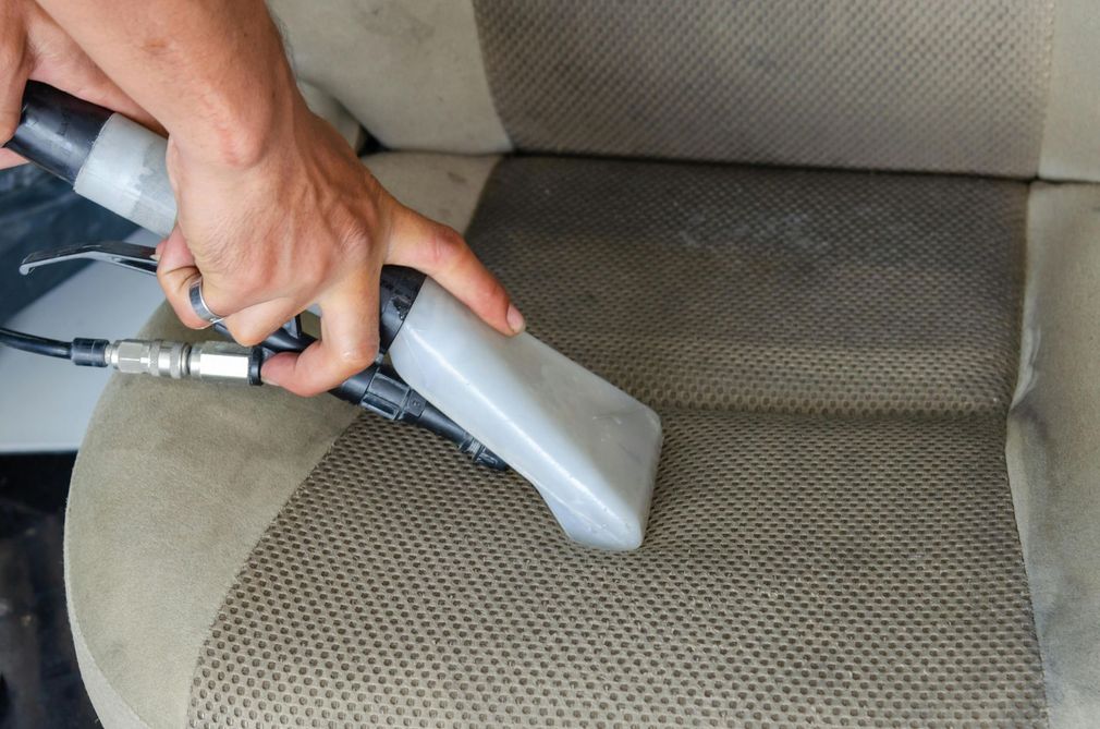 A person uses an upholstery extraction tool to deep clean a beige fabric car seat.