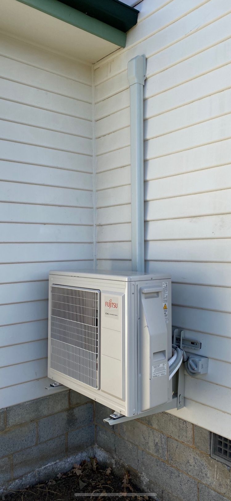 Air conditioner unit mounted on a light-colored building exterior, with metal conduit running up the wall.