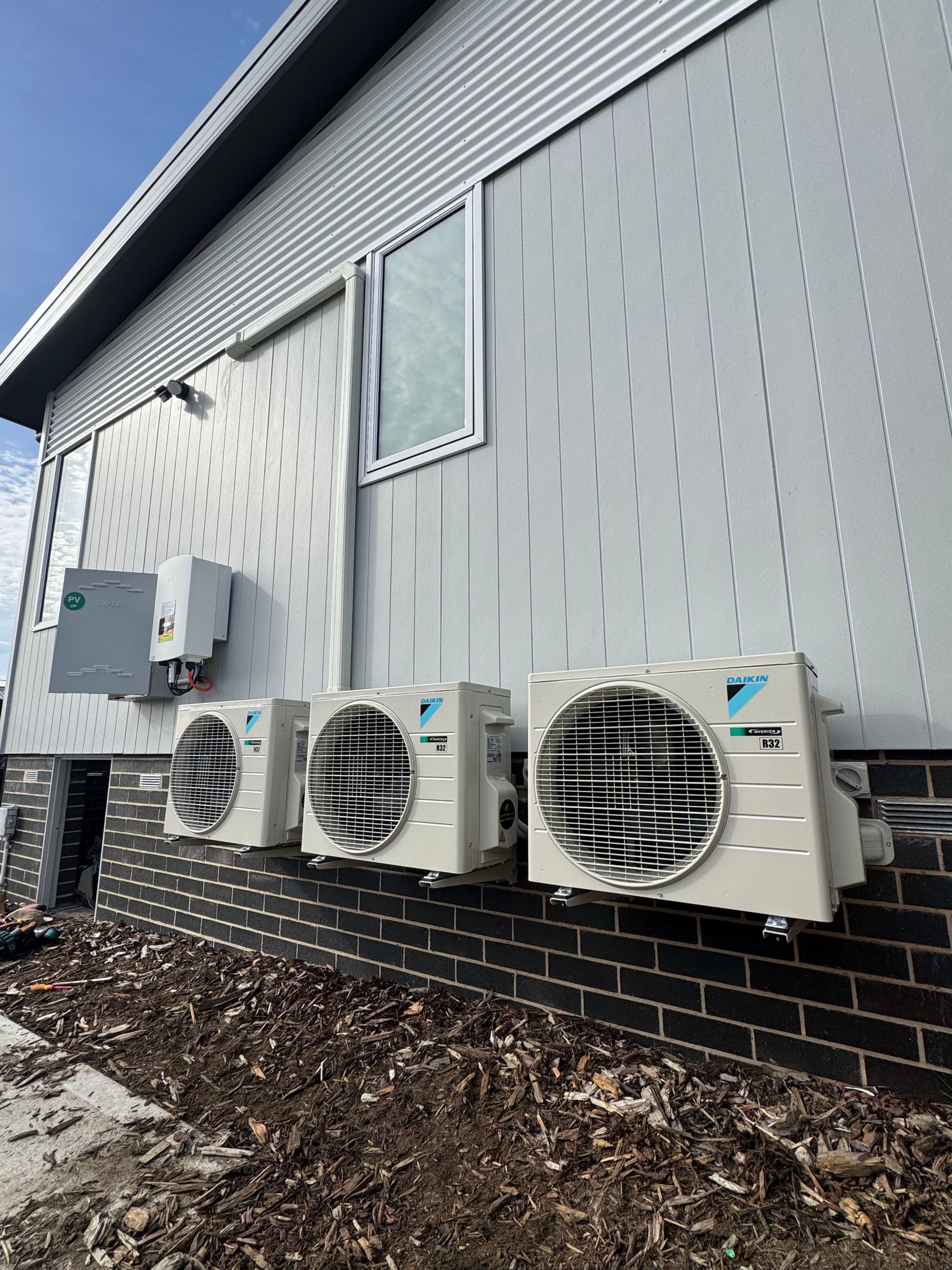 Exterior of a building with three air conditioning units mounted on a gray wall above a brick foundation.