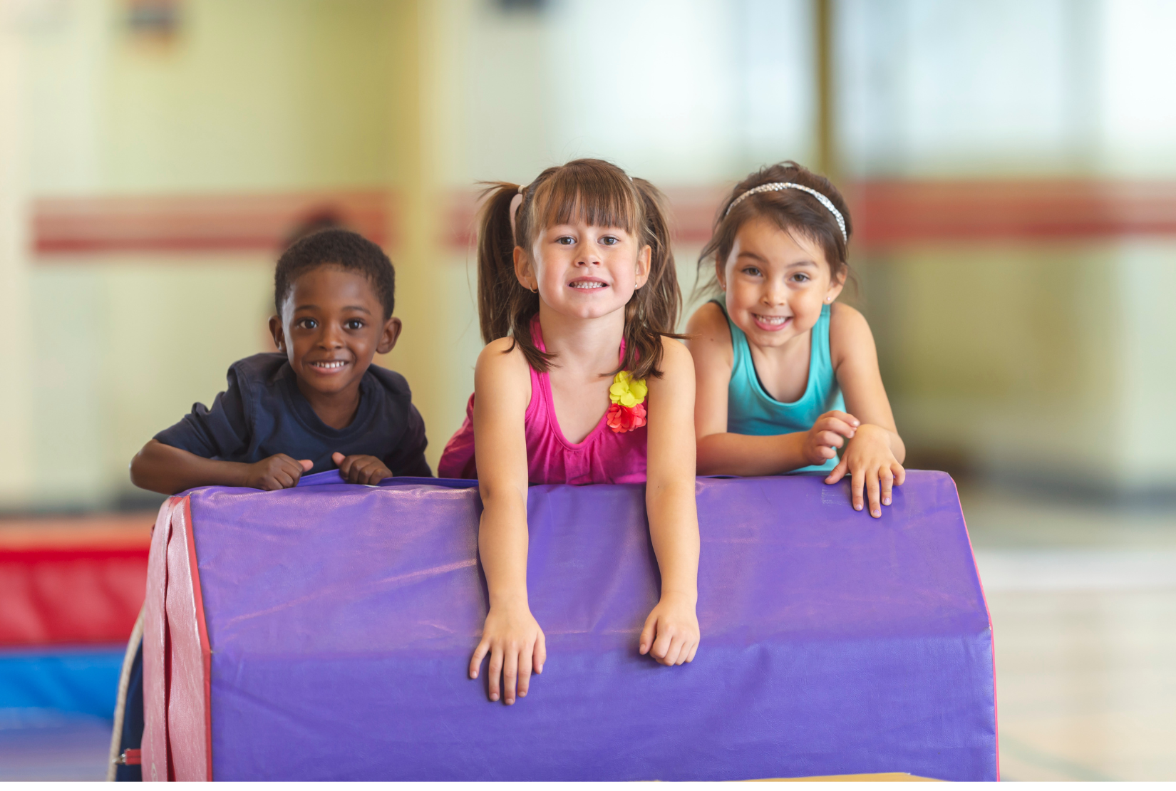 Three children are playing on a purple mat in a gym.