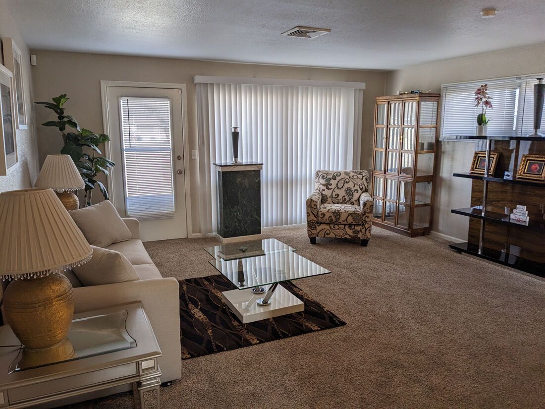 Living room with white sofa, patterned armchair, glass-topped coffee table, and tall display cabinet; window with white curtains.