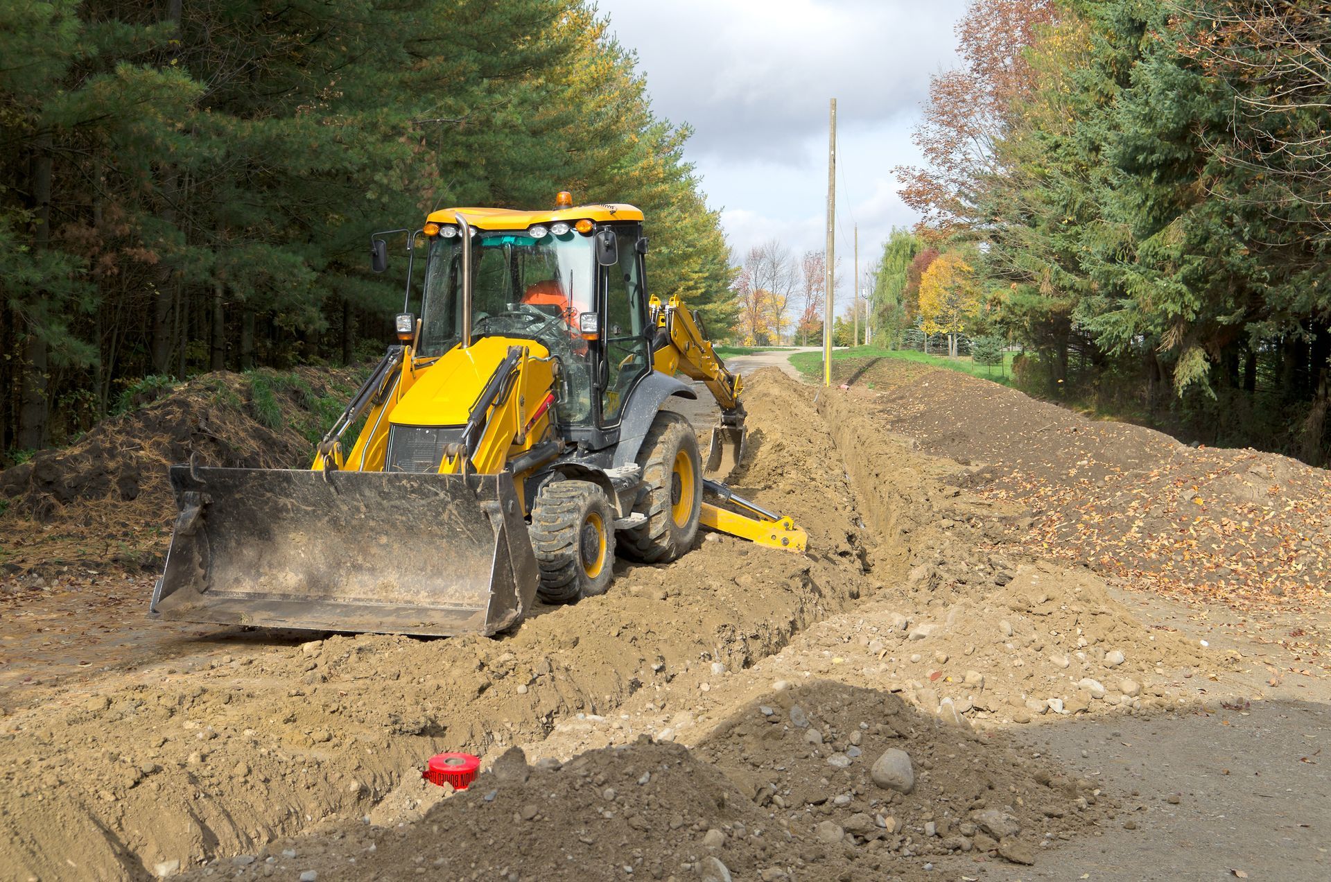 Yellow backhoe digging a trench alongside a road, surrounded by dirt and trees.