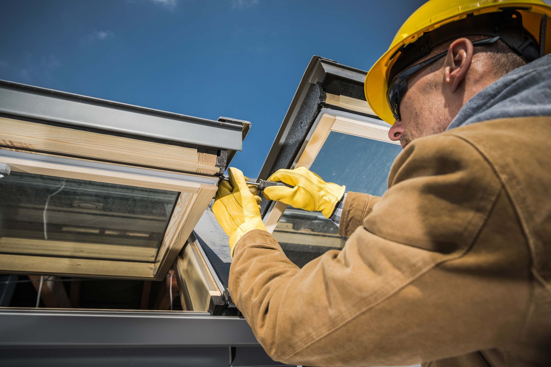 Construction worker in yellow gloves and hard hat, working on a skylight under a blue sky.