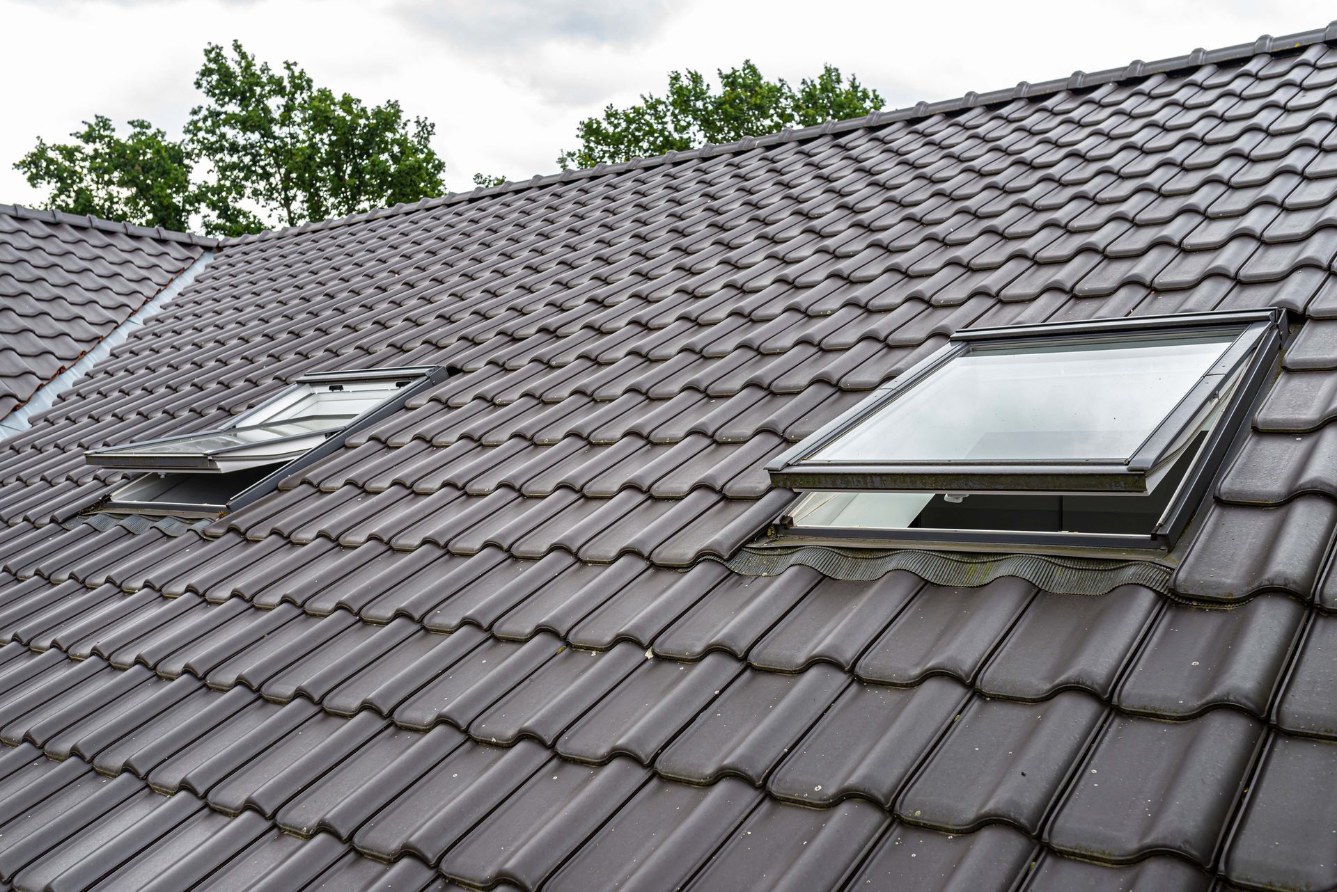 Brown tiled roof with two skylights open against a cloudy sky.